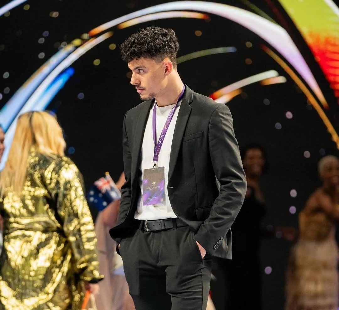 Joven hombre con traje negro y gafete en un evento formal, con fondo colorido y otras personas.