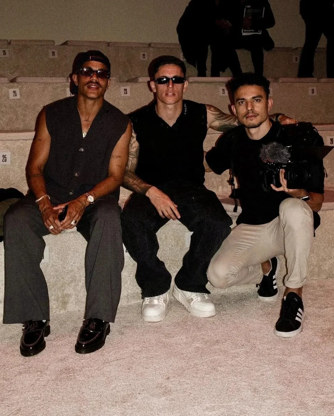 Tres hombres sentados en unas gradas, uno con gafas de sol, otro con tatuajes y una cámara, todos con ropa casual y en un ambiente oscuro.