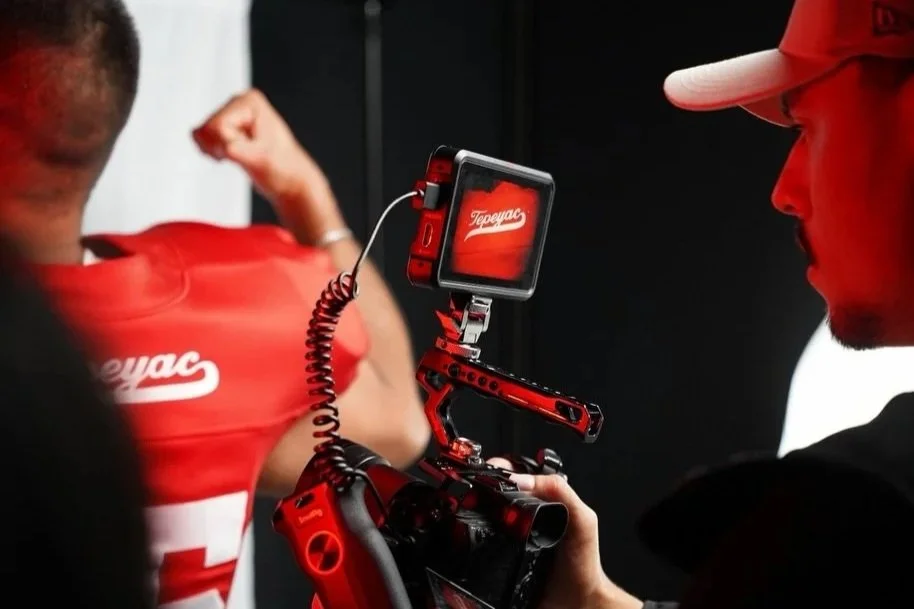 Una persona tomando una fotografía de un jugador de fútbol americano con un uniforme rojo, en un estudio fotográfico, con un fondo negro.