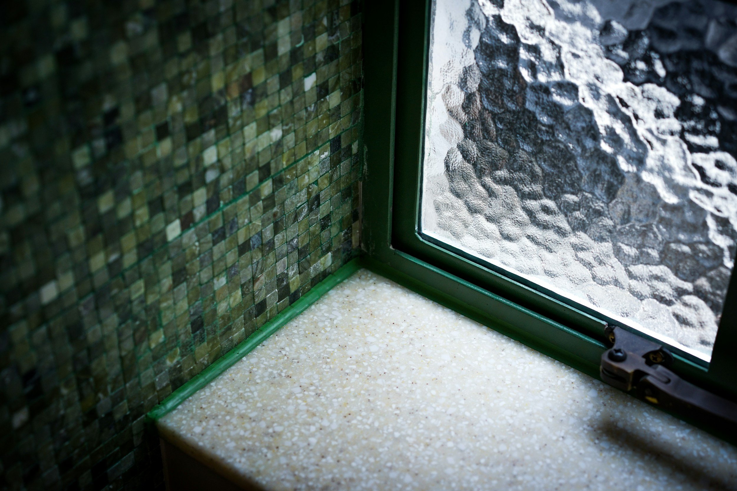 Close-up of a window with textured glass, green frame, and a speckled beige windowsill.