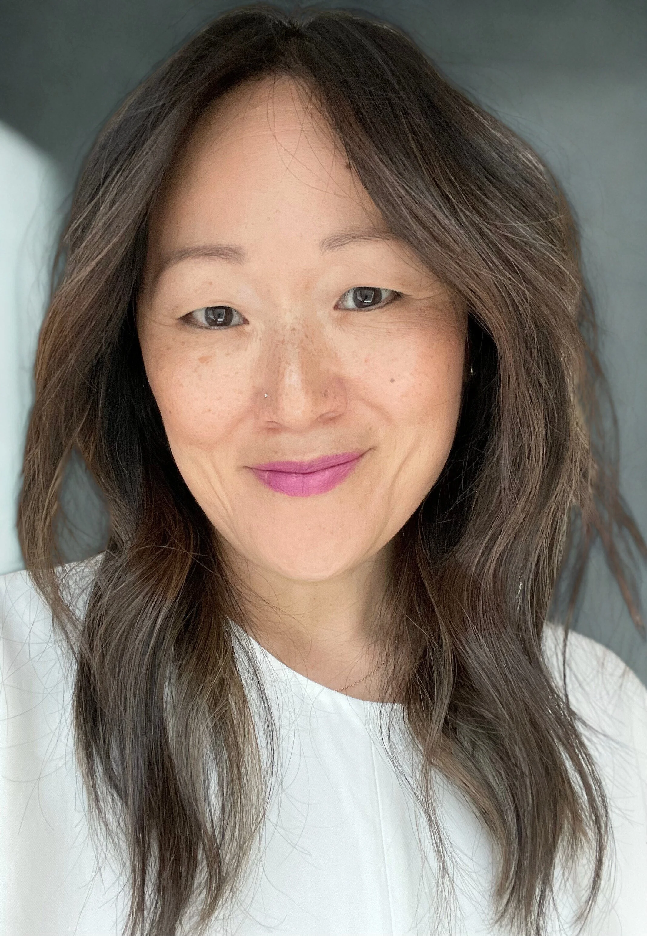 Close-up of a smiling woman with long, wavy brown hair and light makeup, wearing a white top, in a well-lit environment.