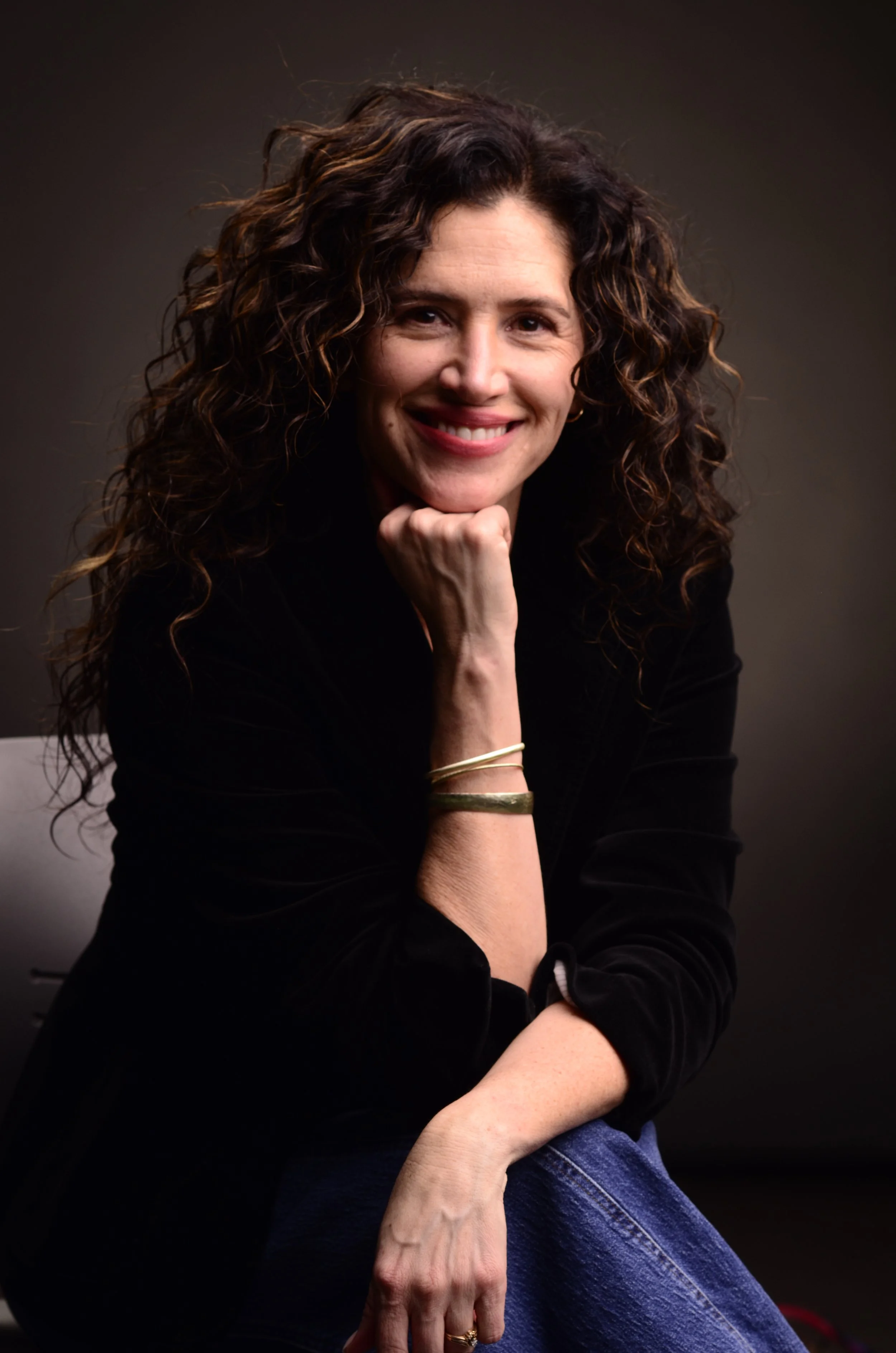 A woman with curly brown hair, smiling, resting her chin on her hand, wearing a black top, blue jeans, and gold bracelets, sitting against a dark background.