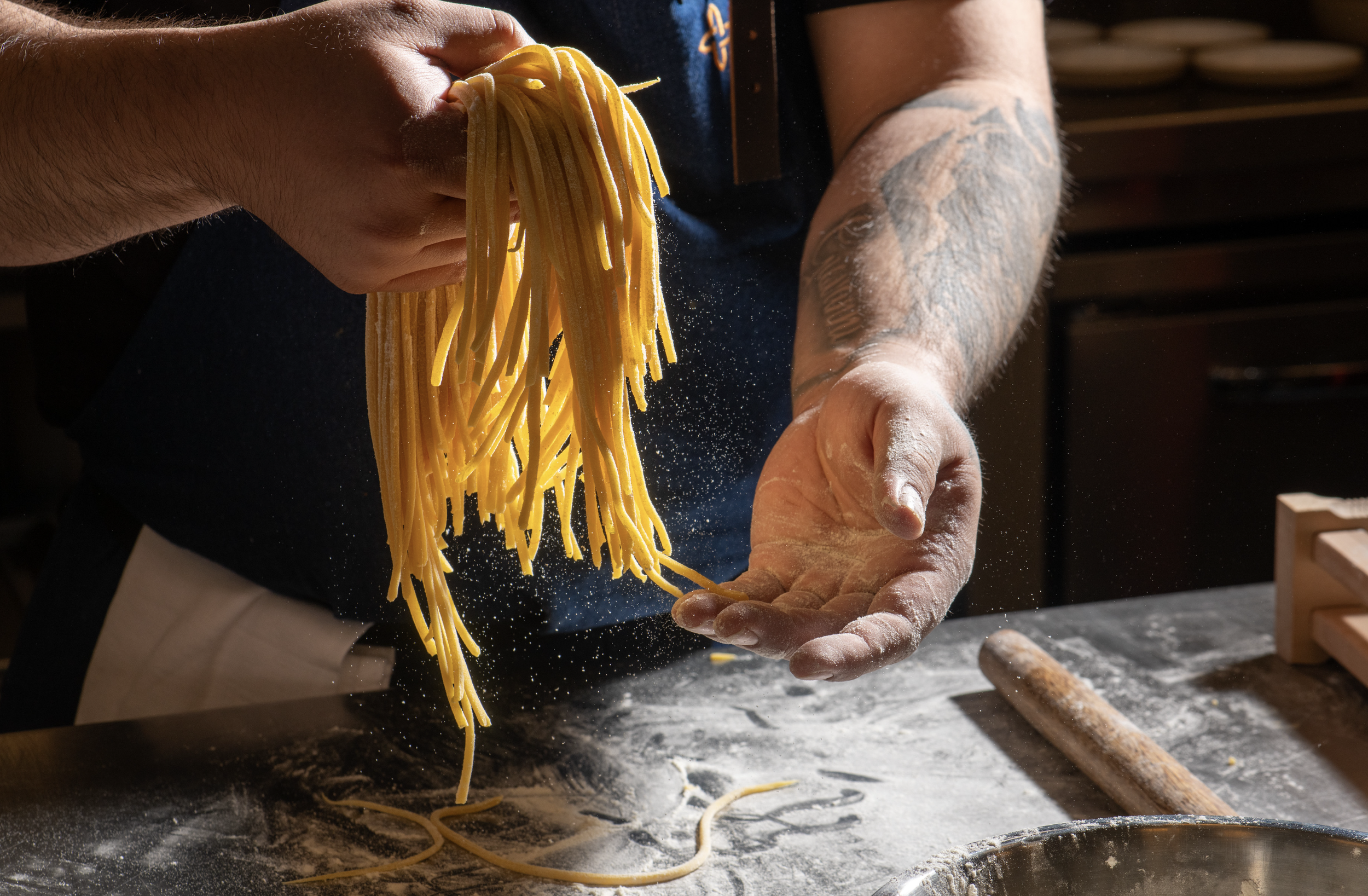 Close-up of a person holding fresh pasta noodles over a floured surface, with a rolling pin nearby and a dark background.