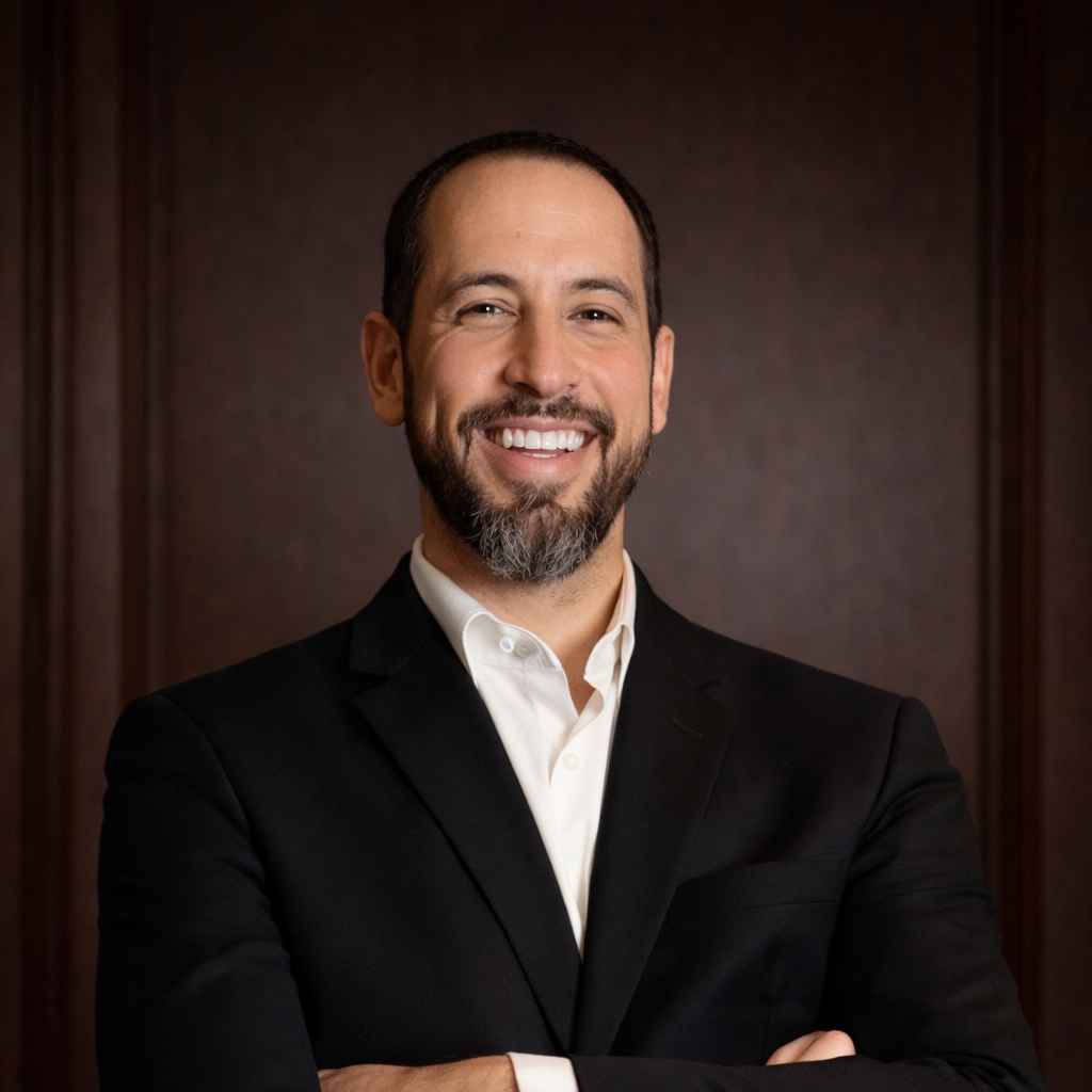 A man with a beard and short dark hair wearing a black suit and white shirt, smiling and standing with arms crossed in front of a dark wood-paneled background.