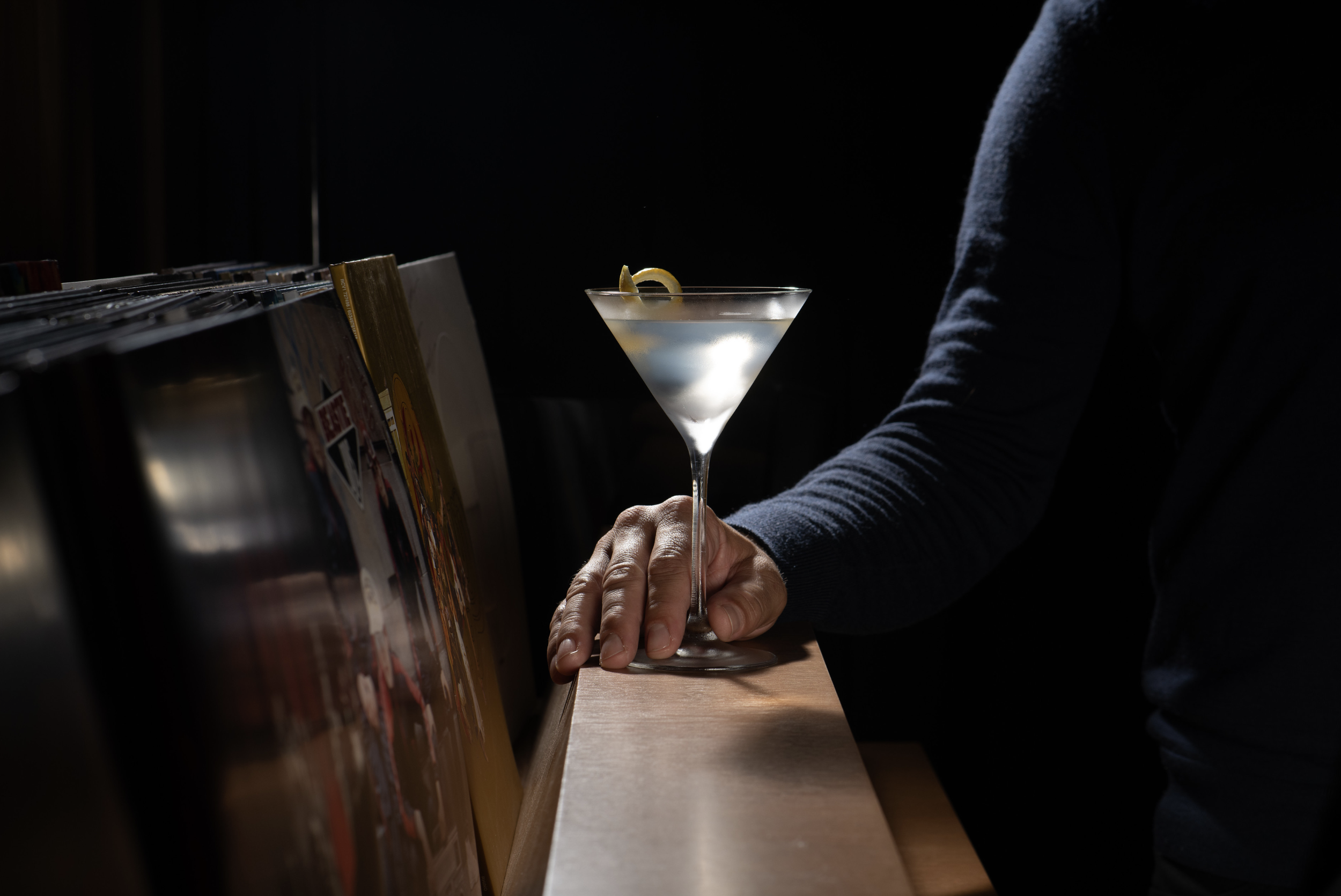 A person selecting a cocktail from a shelf of vinyl records, holding a martini glass with lemon peel garnish in front of their chest.