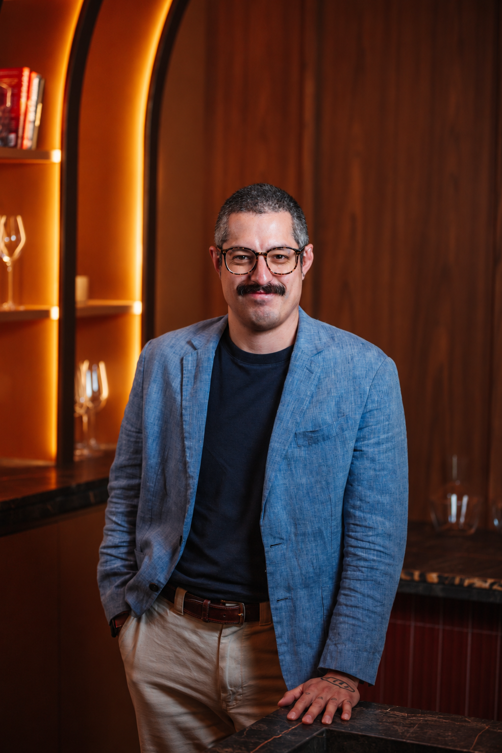 A man with glasses and a mustache holding a cocktail shaker on a bar counter in front of a dark wooden wall.