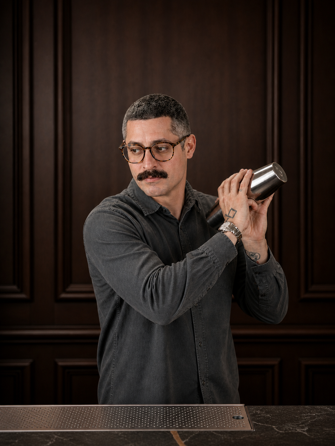 A man with glasses and a mustache holding a cocktail shaker on a bar counter in front of a dark wooden wall.
