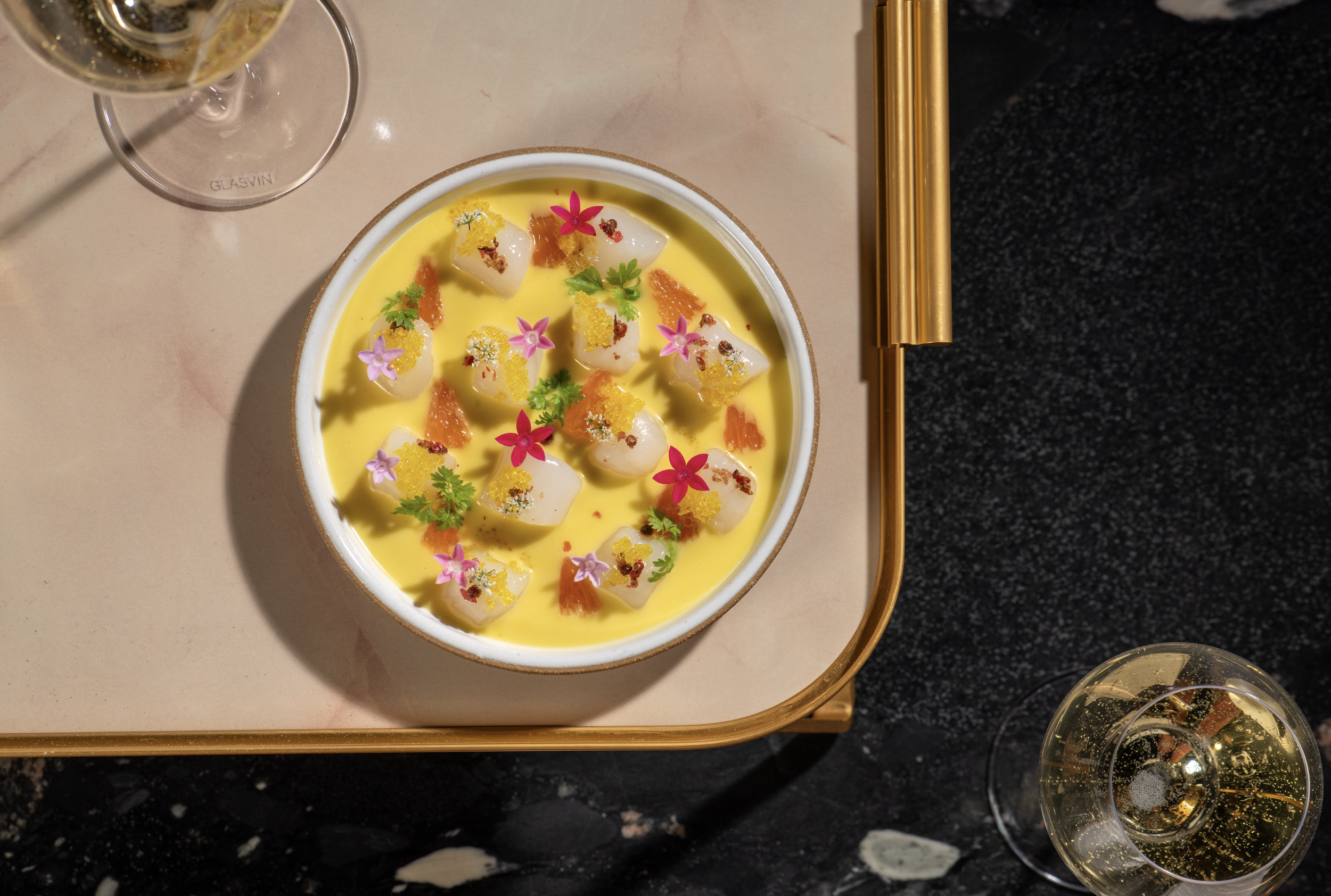 A dessert dish with yellow custard, topped with small pieces of fruit, edible flowers, herbs, and garnishes, served on a white plate. Two glasses of champagne are partially visible, one at the top left and one at the bottom right, on a gold tray with