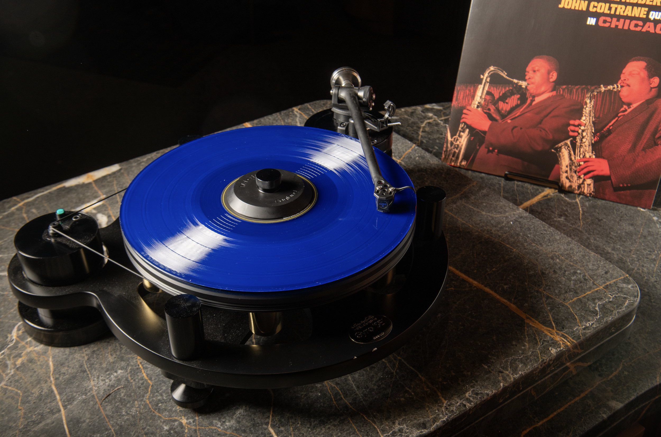 A turntable with a blue vinyl record on it, positioned on a dark marble surface. To the right, there is an album cover featuring two young men playing saxophones.