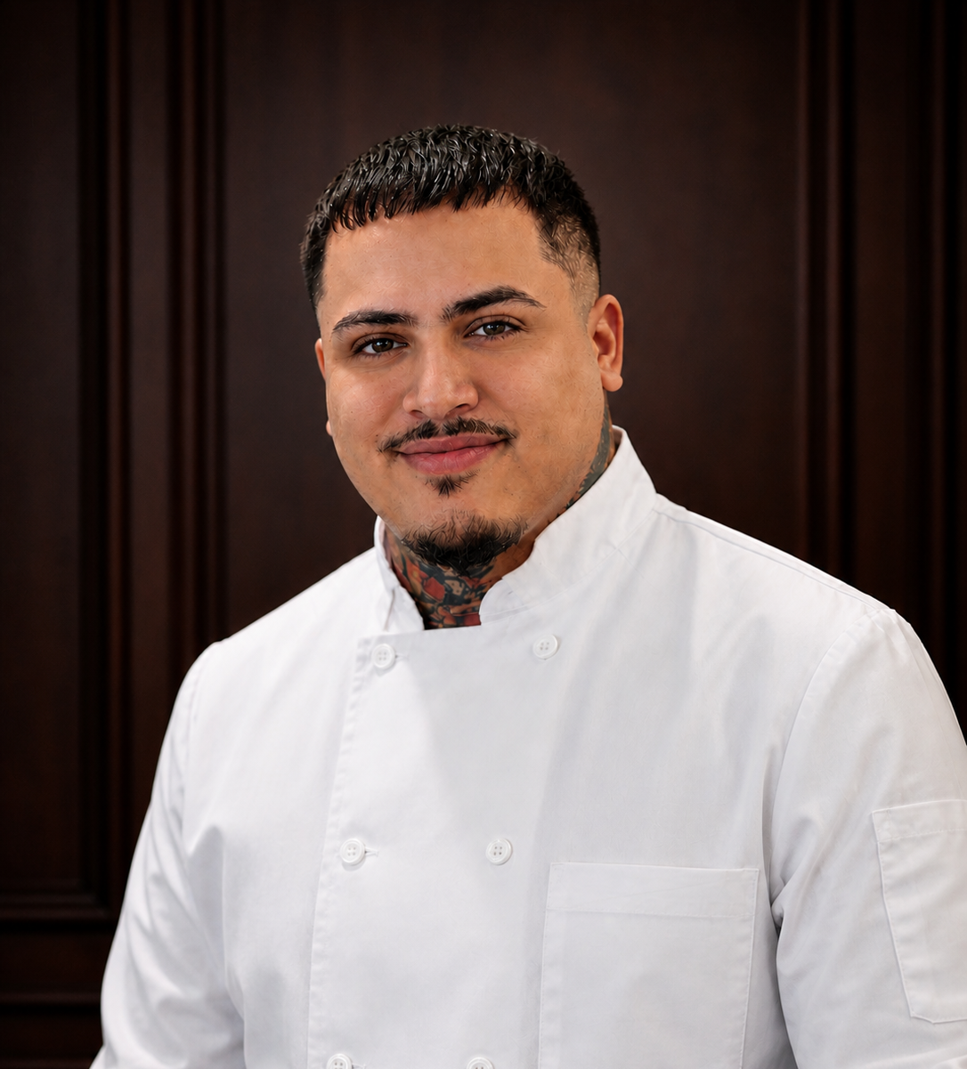 A male chef in a white chef coat standing in front of a dark wooden background.