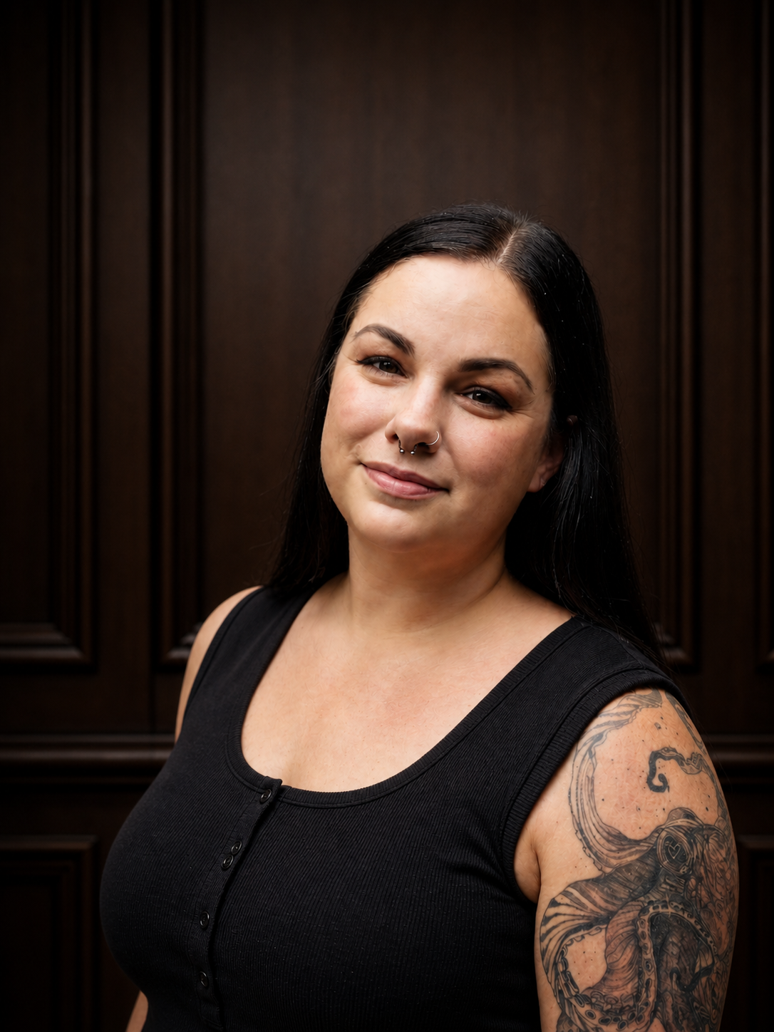 Portrait of a woman with black hair, tattoos on her left arm, and a septum piercing, wearing a black sleeveless top, standing against a dark wooden paneled background.
