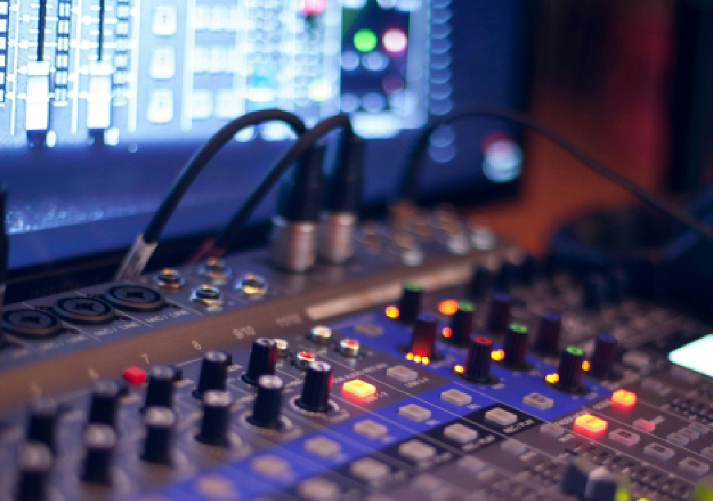 Mixer audio con pulsanti e leve, schermo digitale sullo sfondo in un ambiente di registrazione o DJ.