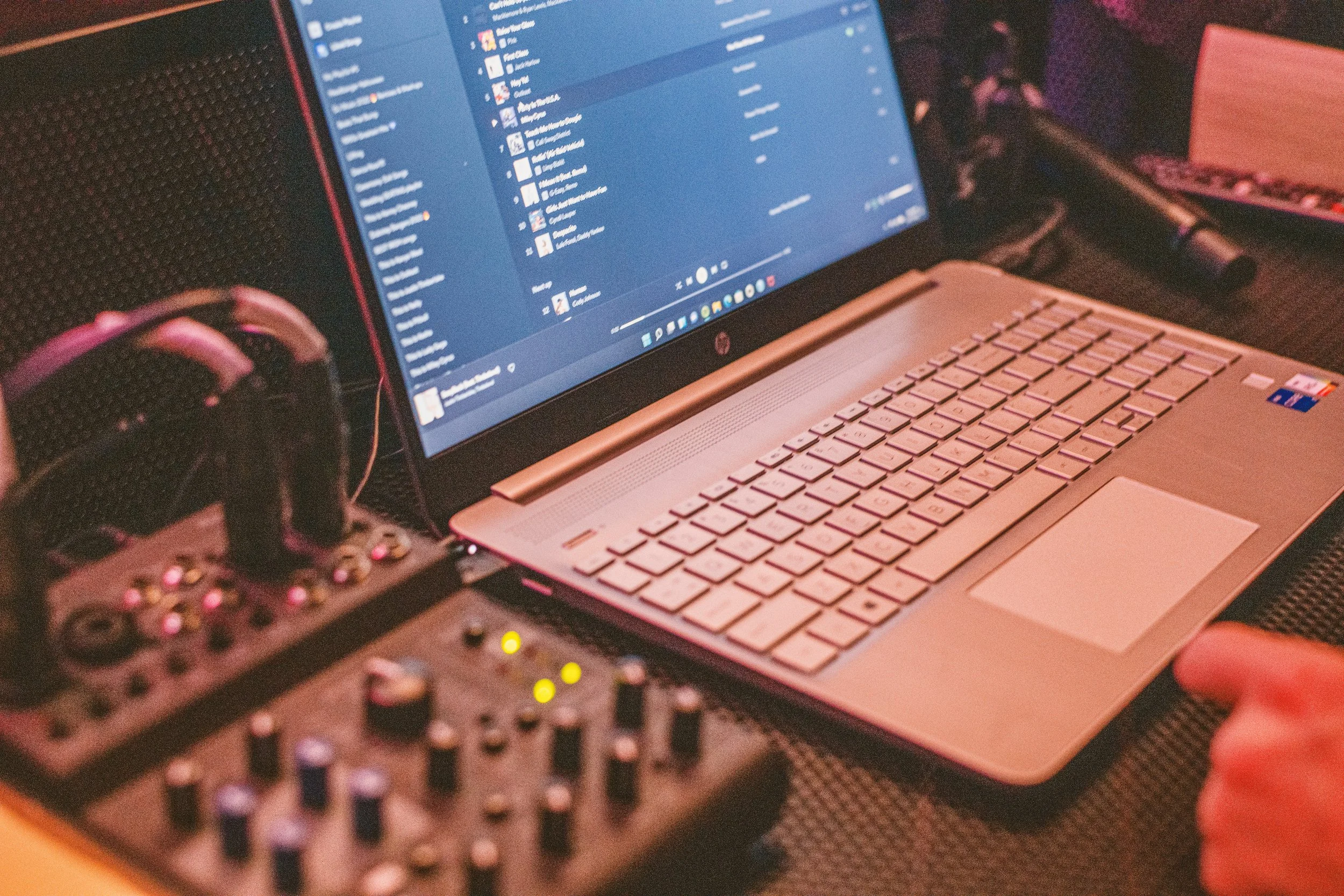 Laptop aperto con schermo con software musicale o di produzione audio, mixer audio con manopole e luci, cuffie sospese, e una mano che indica una determinata zona del laptop.