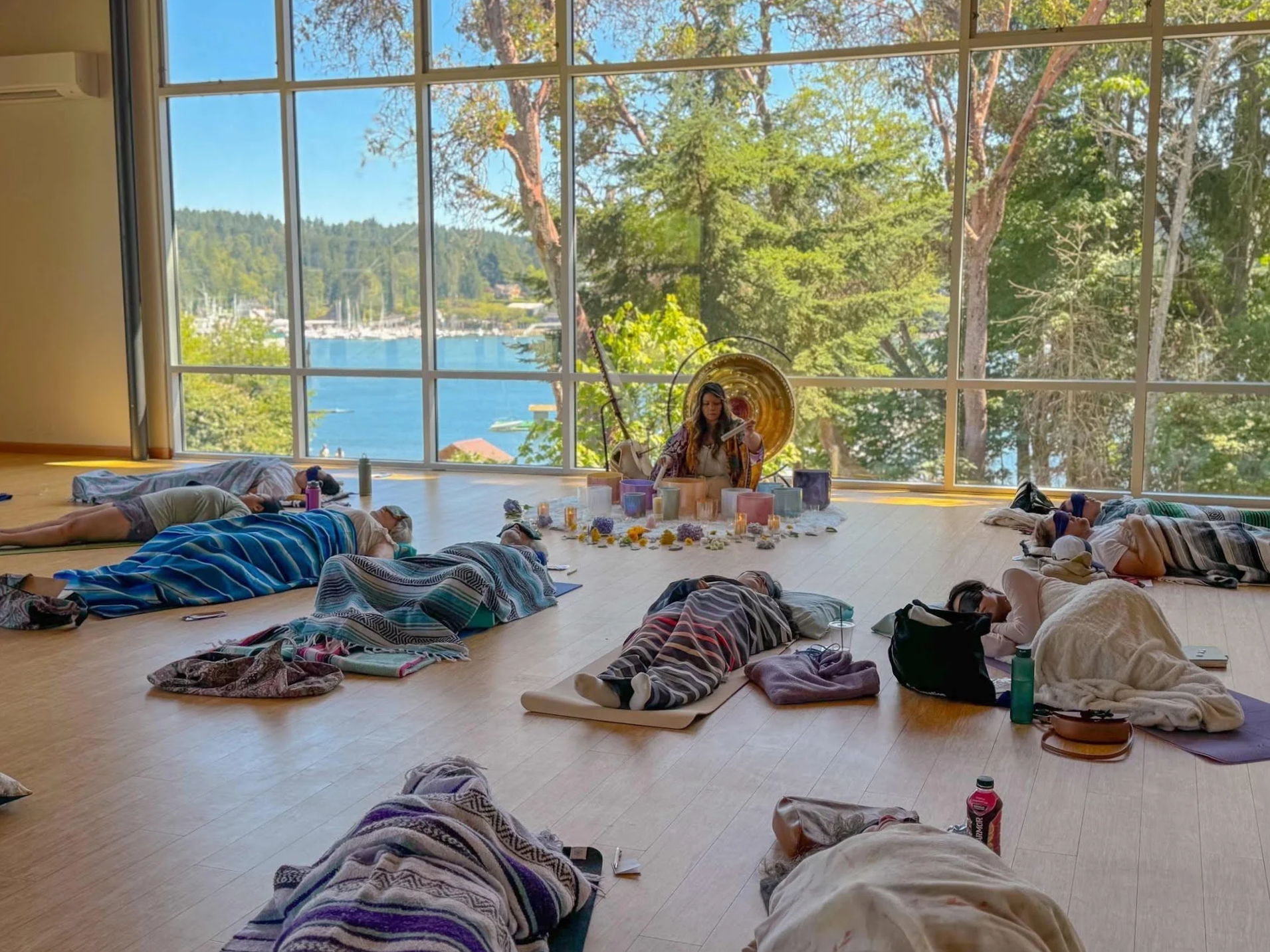 A group of people lying on the floor on yoga mats during a meditation session in a spacious room with large windows showing trees and a body of water outside. A woman is sitting in front, leading the session, with various objects including candles and flowers on a table in front of her.
