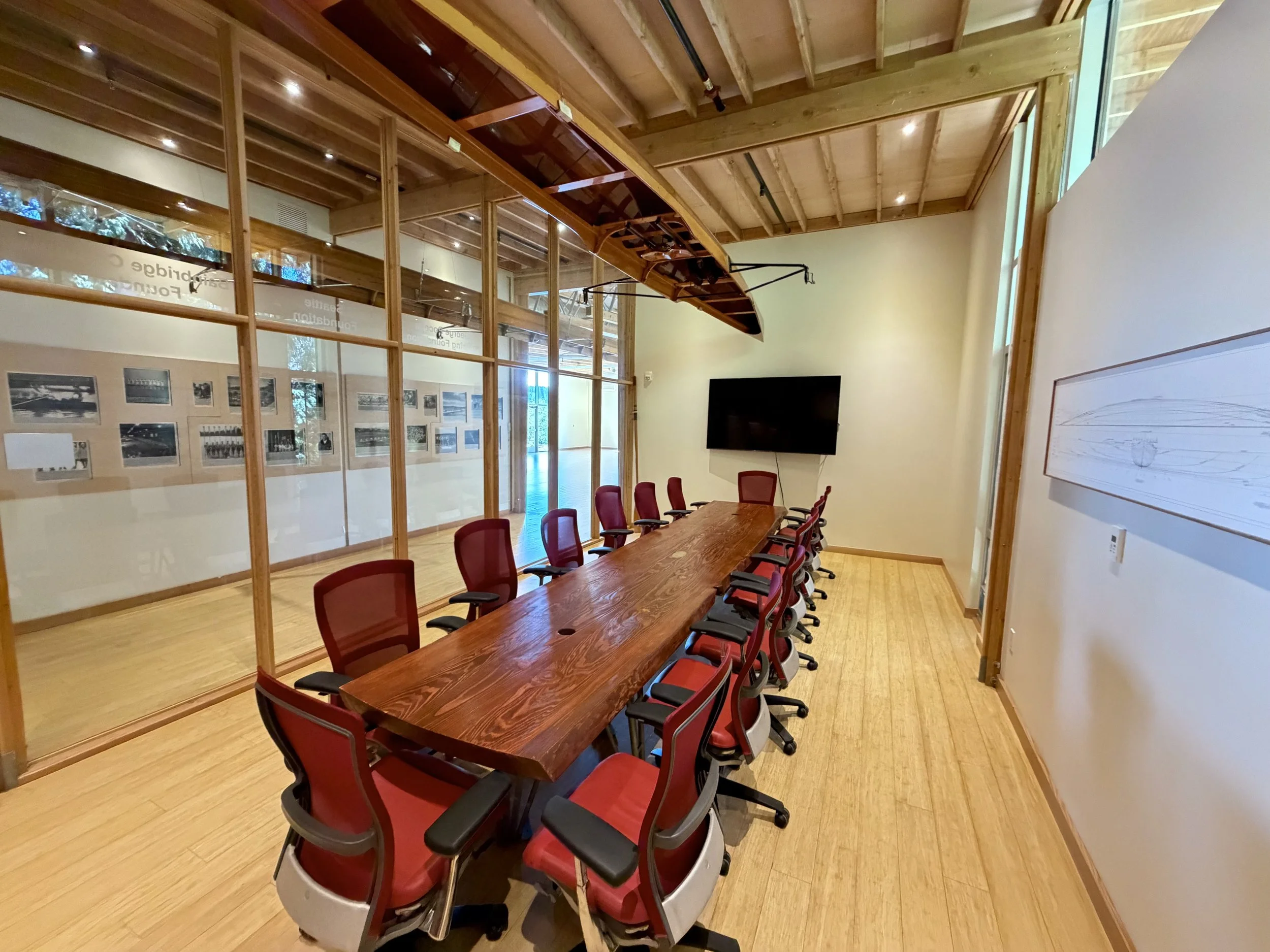 A conference room with a long wooden table surrounded by red and black office chairs. There is a large mounted flat-screen TV on the wall, and a canoe hanging from the ceiling. The room has wooden beams on the ceiling and a glass wall with history photographs displayed on the adjacent wall.