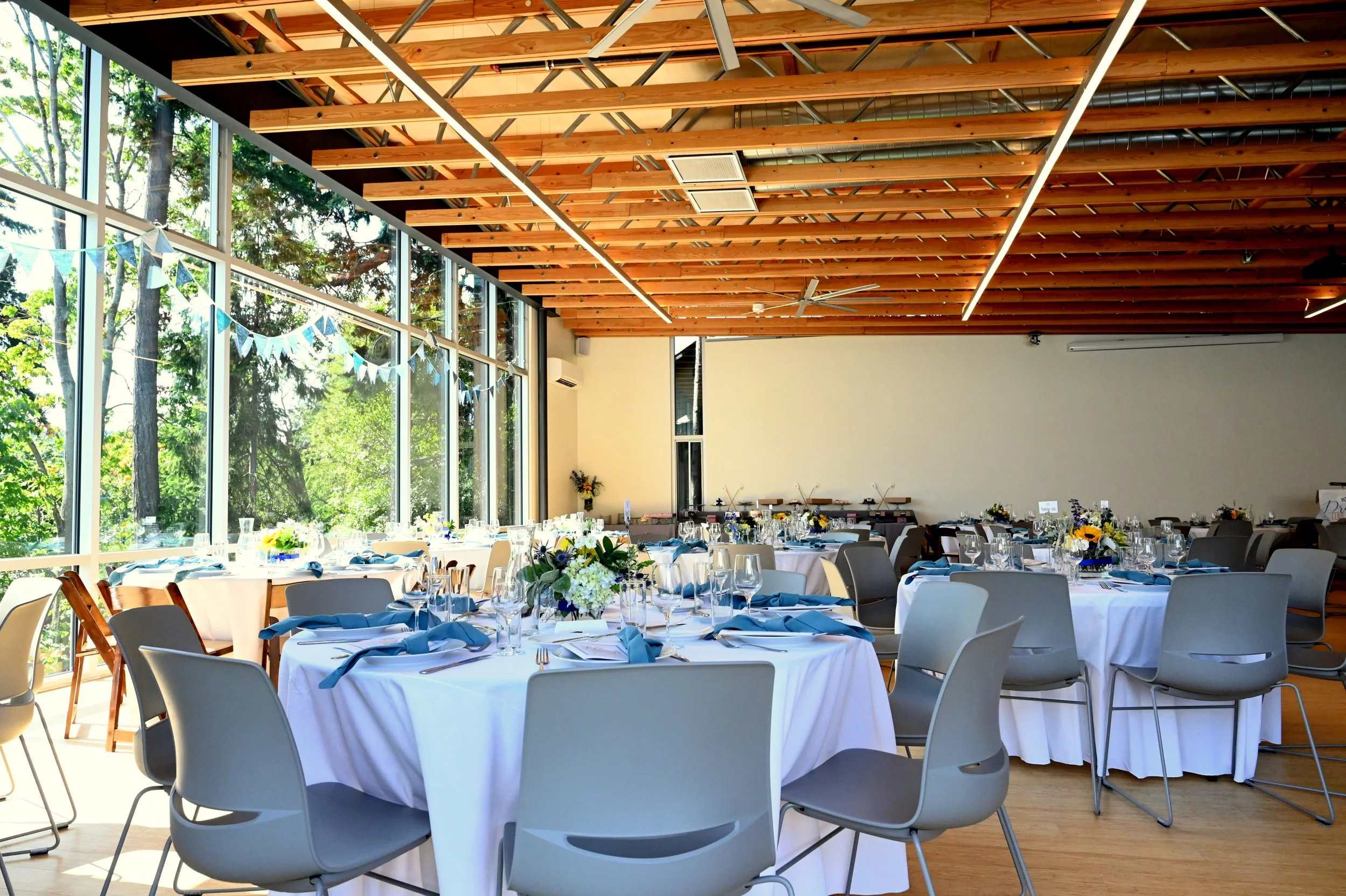 A banquet hall with decorated tables for a celebration, large windows showing trees outside, and a high wooden ceiling with light fixtures and ceiling fans.
