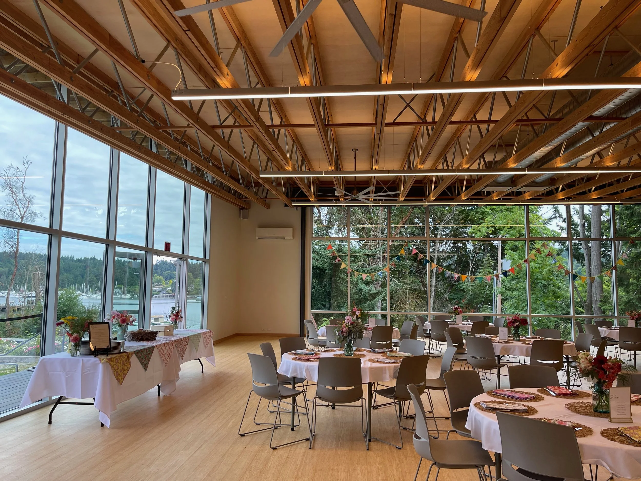 An indoor venue with large windows and a view of trees and water outside, decorated for a celebration with round tables covered in white tablecloths, floral centerpieces, colorful bunting, and place settings, with a wooden ceiling and hanging lights.