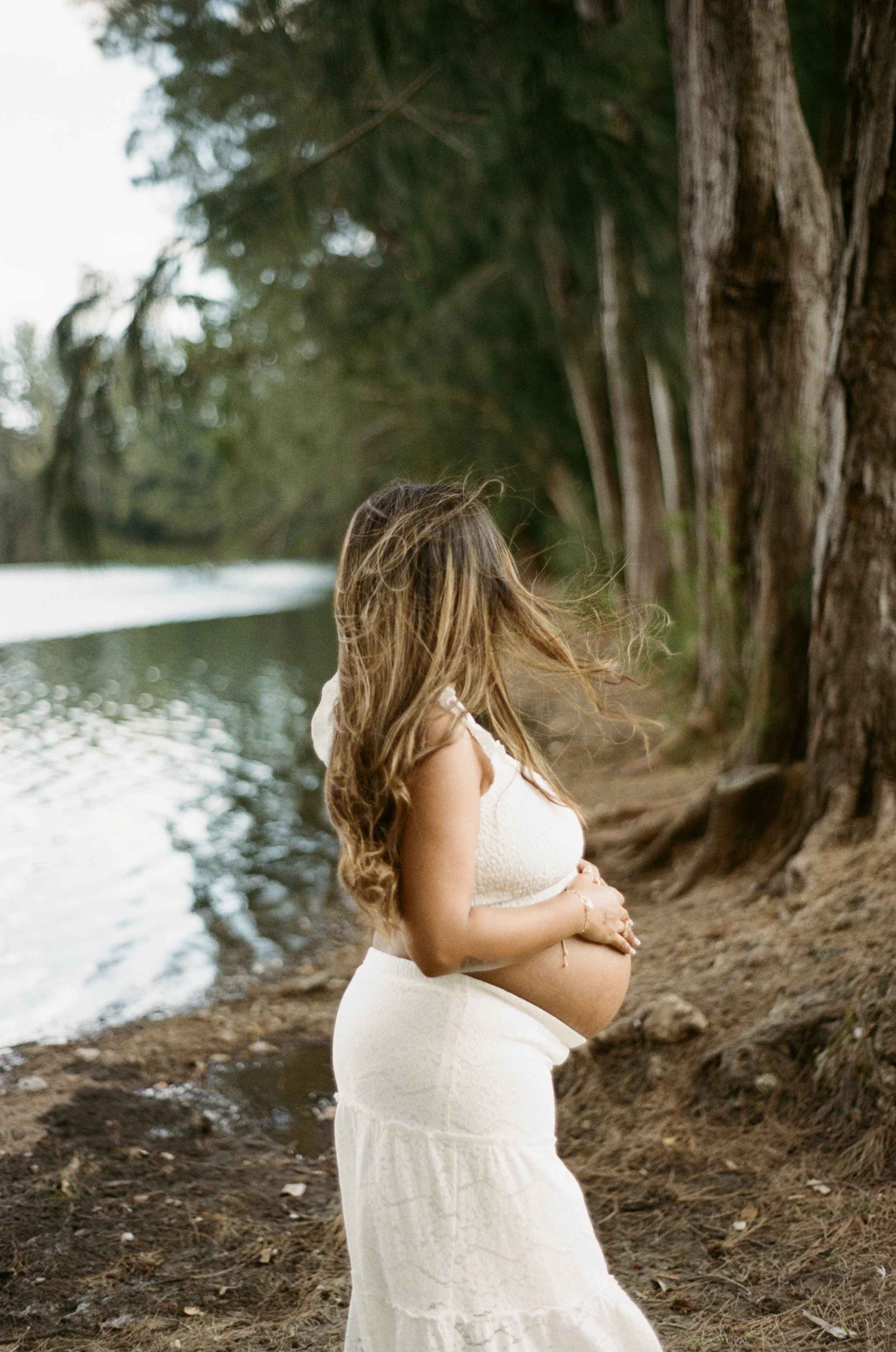 A pregnant woman with long wavy hair, dressed in a white sleeveless dress, stands by a riverbank with her hands on her pregnant belly, surrounded by tall trees and natural scenery in Davie, Florida.