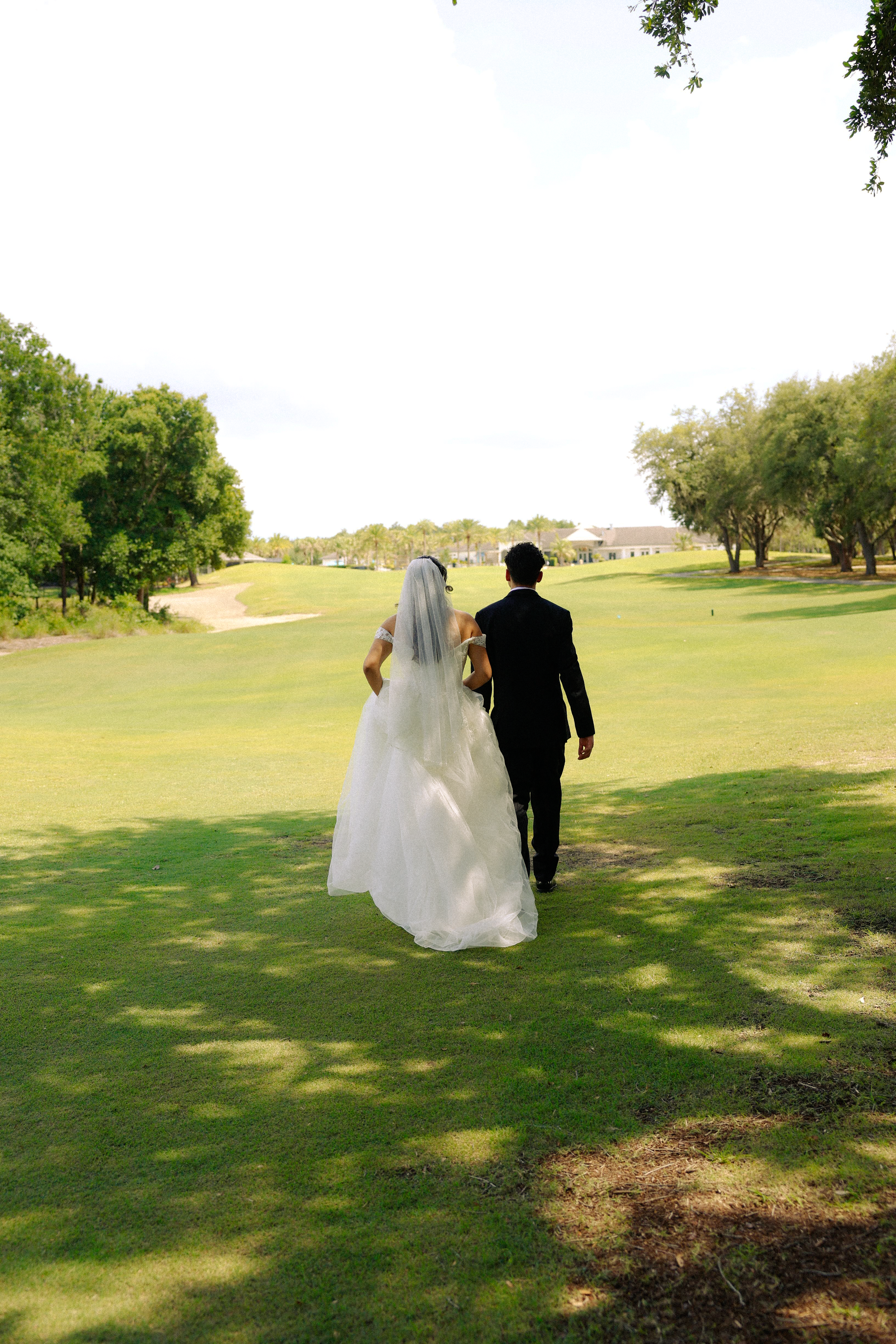 A bride and groom walking together on a lush green golf in Orlando, Florida course during daytime, surrounded by trees and open sky.