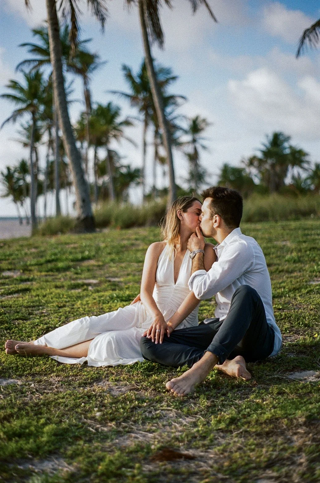 A couple sitting on grass, sharing a kiss, surrounded by palm trees on a beach during daytime.