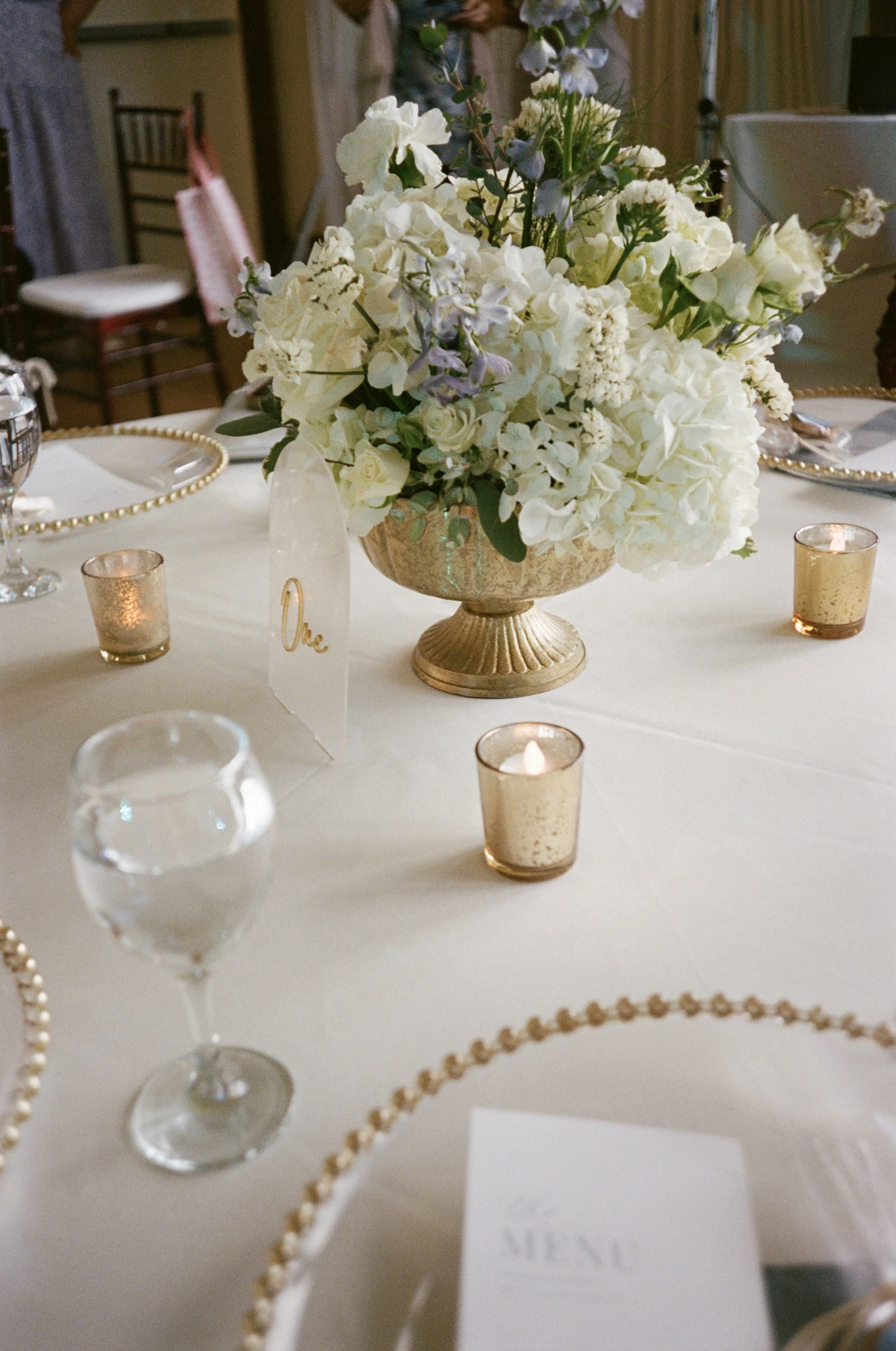 A large floral arrangement with white flowers in a decorative gold vase on a banquet table, surrounded by candles, glasses, and place settings in Davie, Florida.