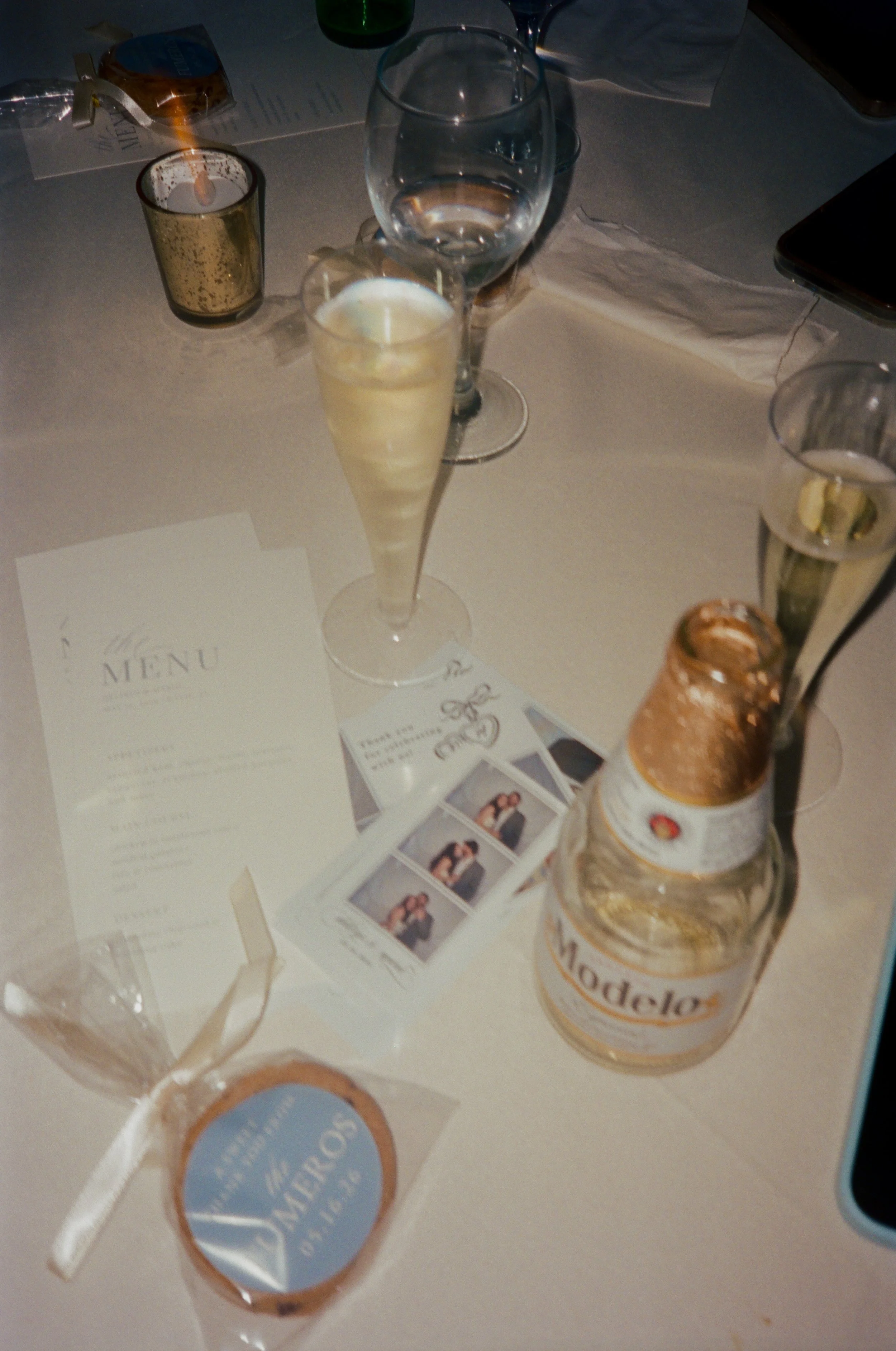 A table with various drink glasses, including champagne flutes and a beer bottle, along with some paper menus and printed photos. There is a lit candle in a gold holder and a smaller candle on the table in Davie, Florida.