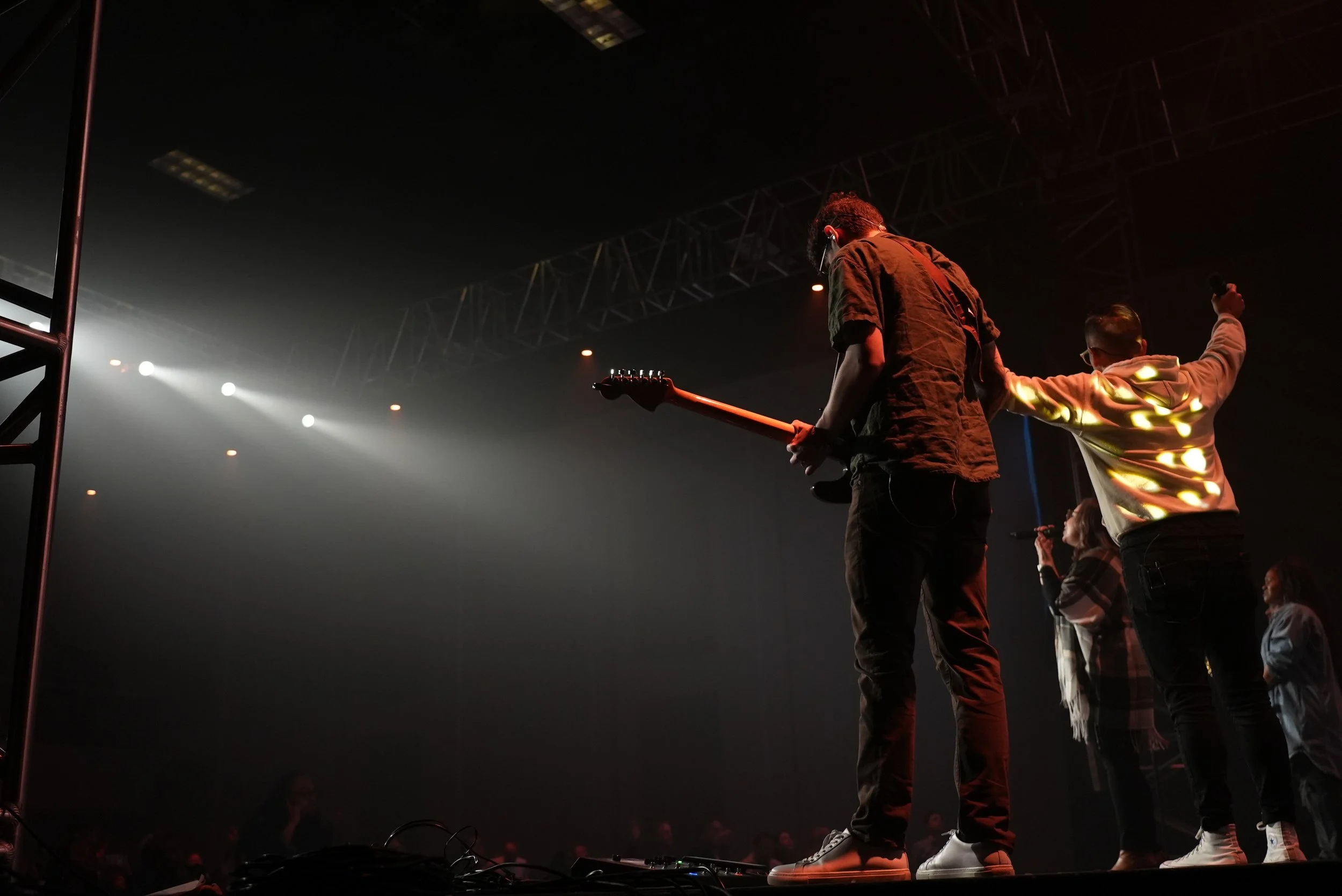 Two musicians on stage, one playing guitar with head bowed, the other raising a hand, surrounded by stage lighting and audience.