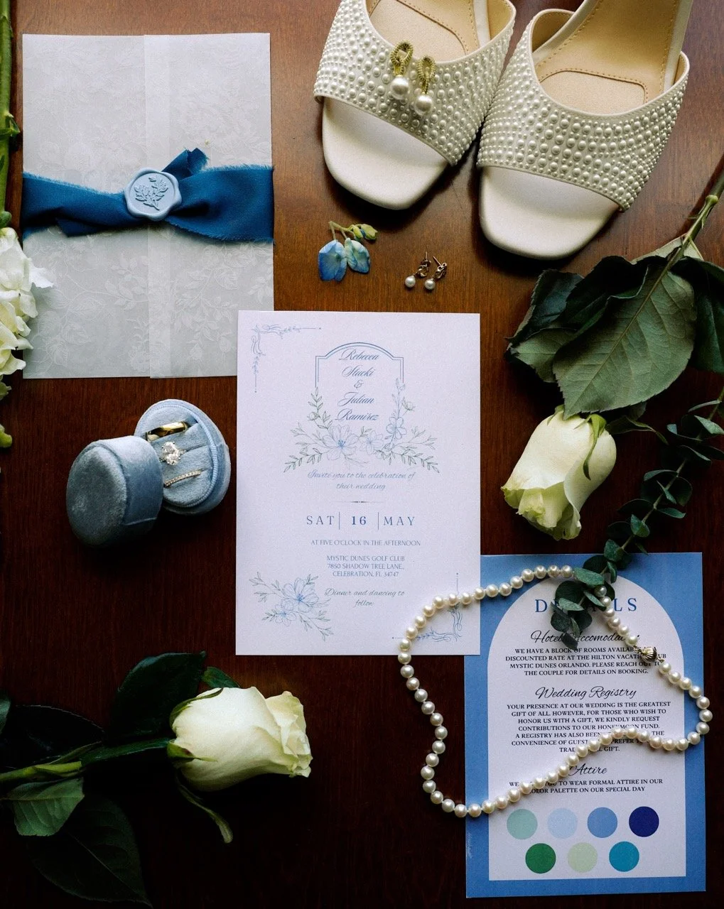 Detail shots with a Wedding invitation, pearl jewelry, white wedding shoes with pearl embellishments, white roses, blue ribbon with wax seal, earrings, and wedding rings displayed on a wooden surface in Orlando, Florida.