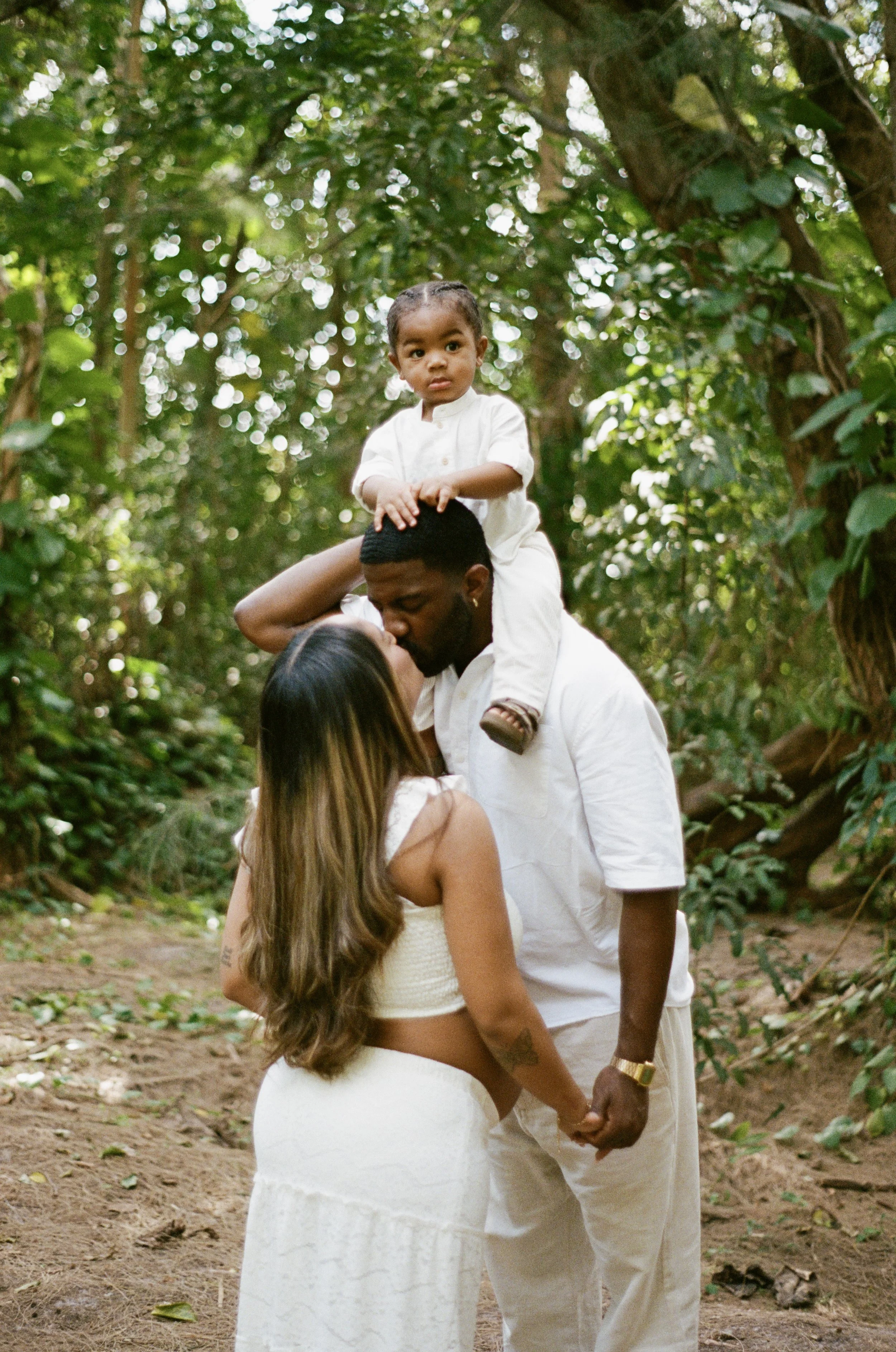 A couple holding hands and kissing outdoors in a forest, with a young boy sitting on the man's shoulders, looking directly at the camera in Florida.