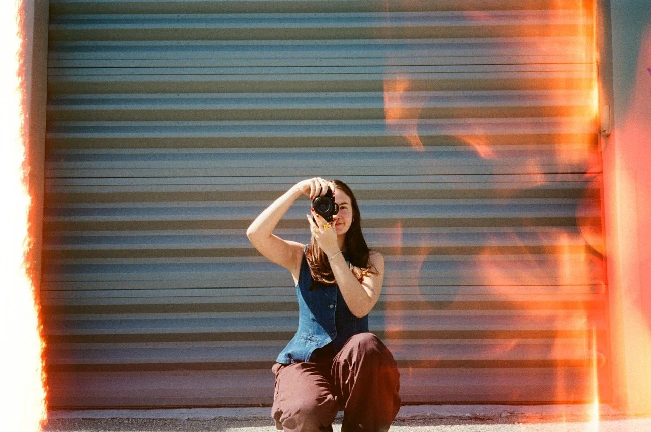 Young woman taking a photo with a camera in front of a corrugated metal wall, with orange light leaks on the sides.