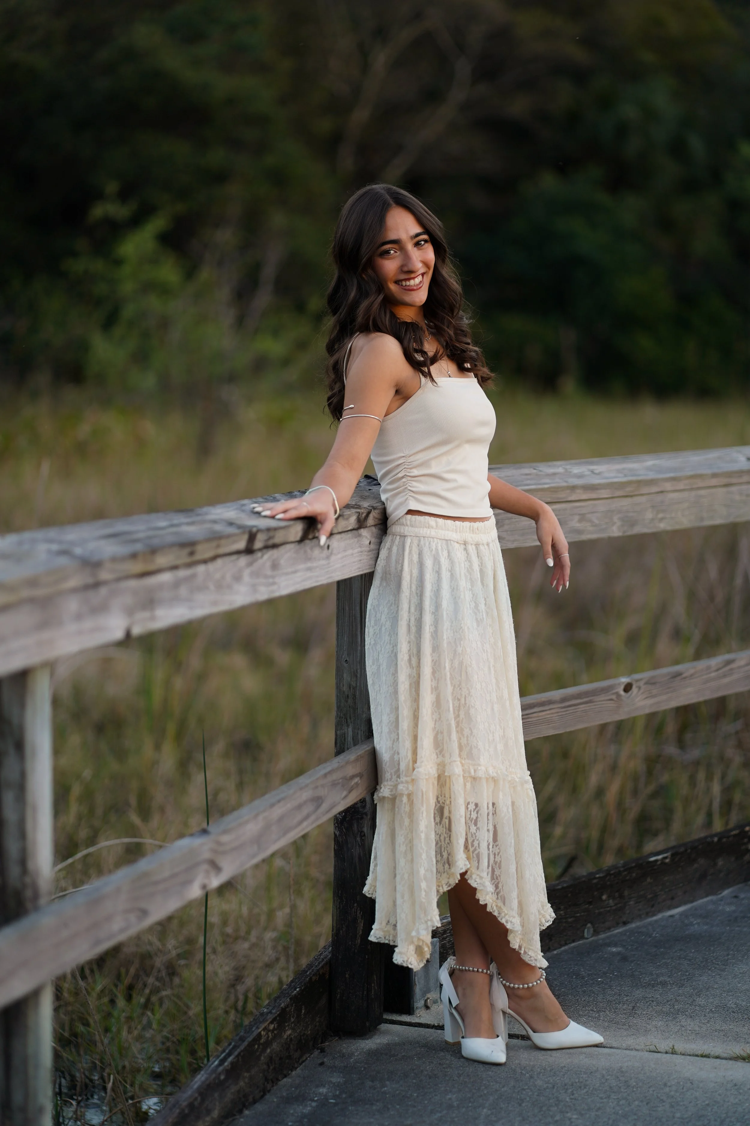 A young woman in a cream-colored lace dress and white heels with pearl ankle straps, smiling and leaning on a wooden railing outdoors with green trees in the background.