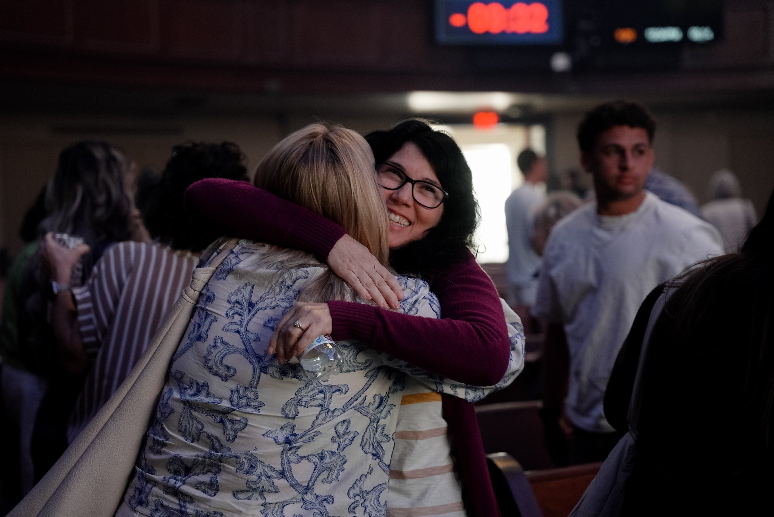 Two women hugging and smiling in a crowded indoor setting, with one wearing glasses and the other holding a water bottle.