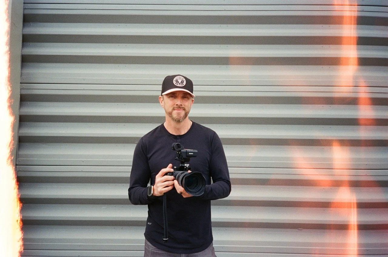 A man holding a professional camera with a flash attachment, standing in front of a gray corrugated metal wall, wearing a black long-sleeve shirt and a black cap.