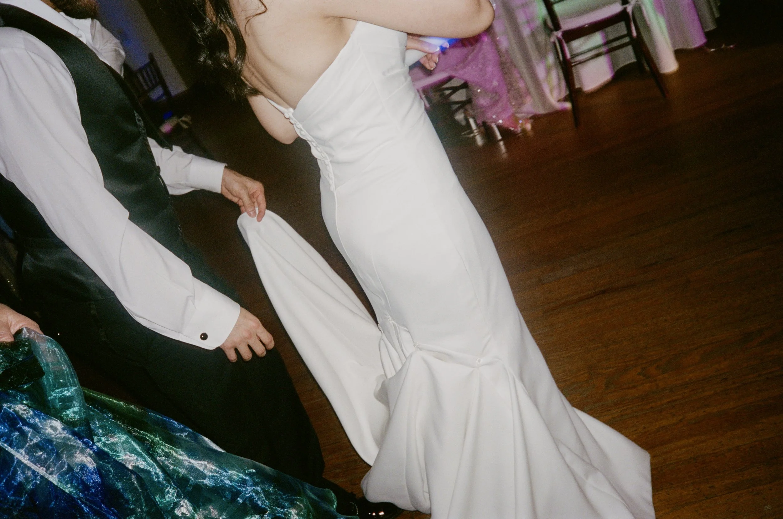 A woman in a white wedding dress standing indoors at a reception, with chairs and tables in the background.