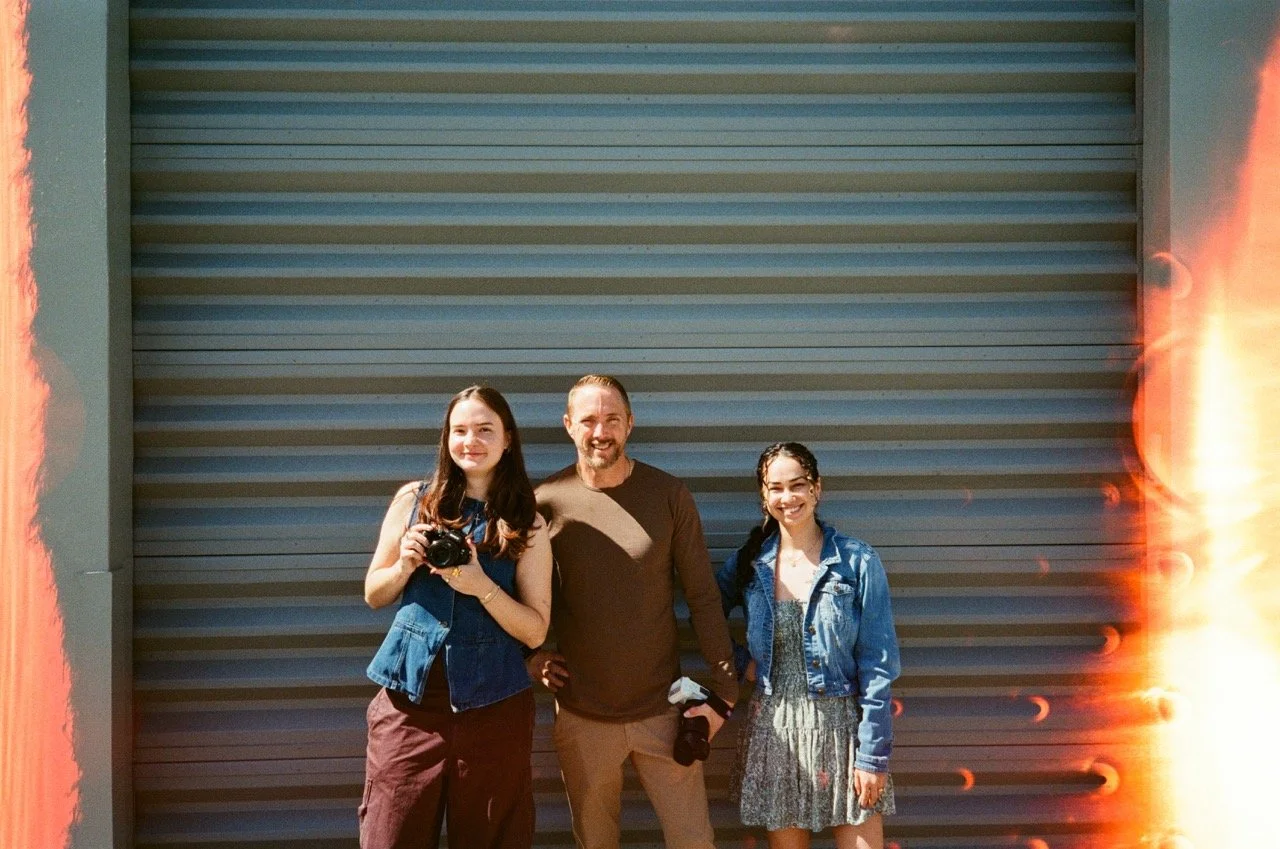 Three people standing in front of a large industrial door with flames on both sides, holding cameras and smiling.
