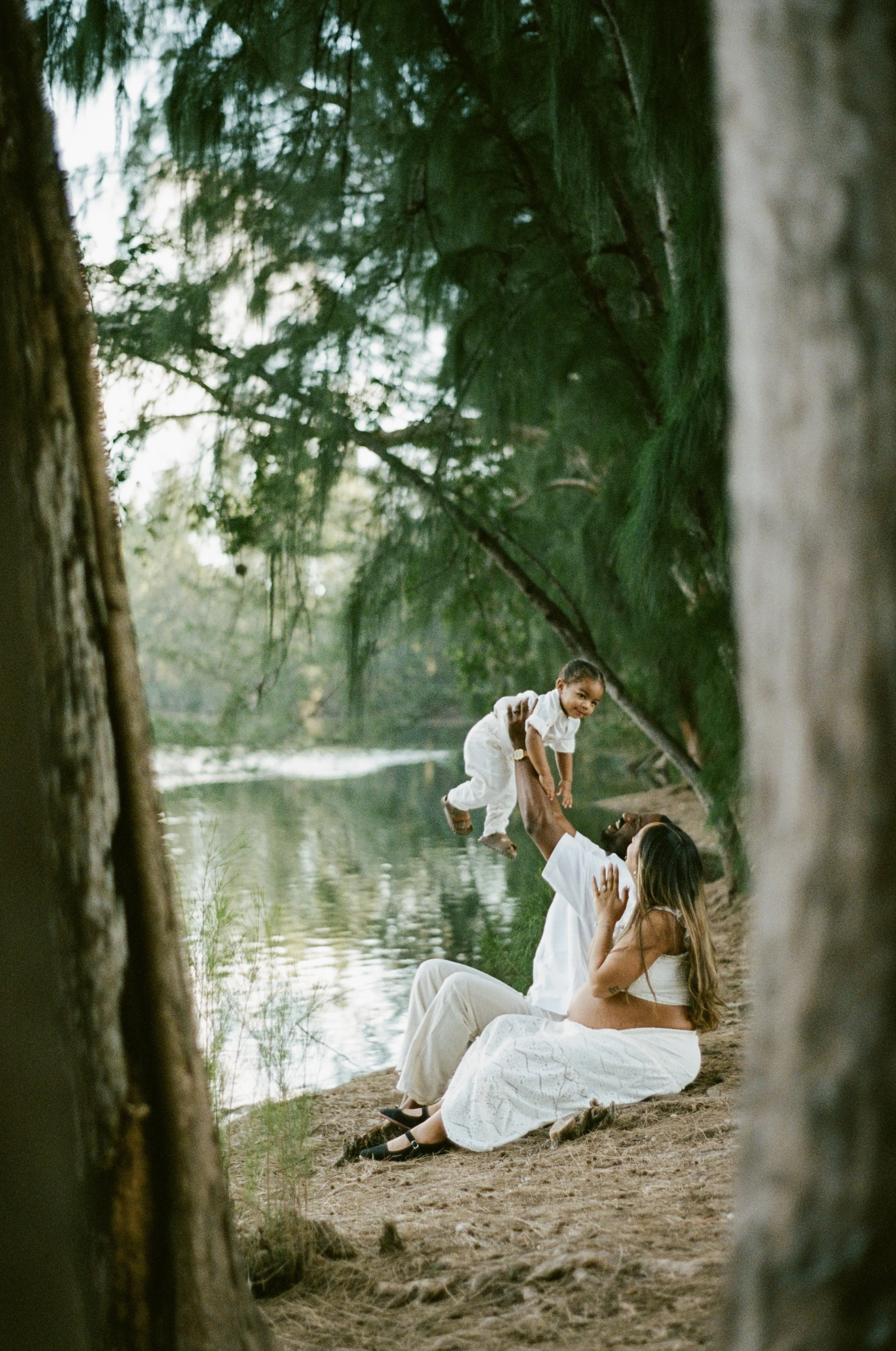 A woman and a man sitting by a river, playing with a young child who is jumping into the woman's arms, surrounded by trees and greenery.