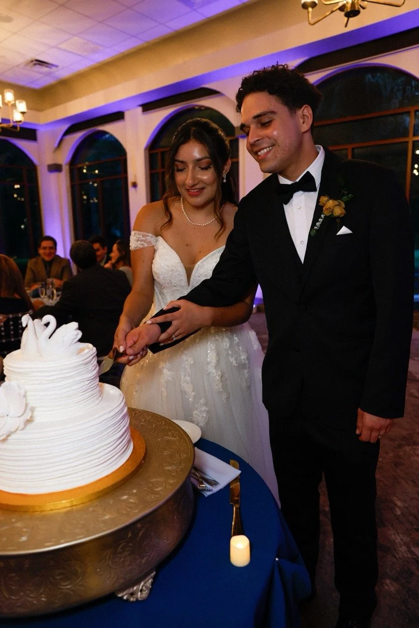 Bride and groom cutting a wedding cake with swan-shaped decorations, indoors with guests seated at tables in the background.