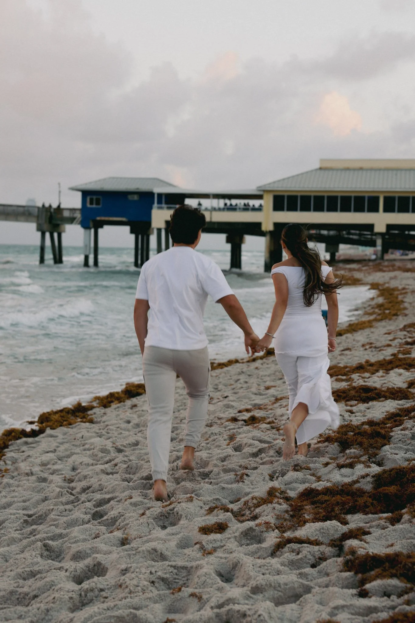 A couple walking hand in hand on a sandy beach near the ocean, with pier buildings in the background and cloudy sky overhead.
