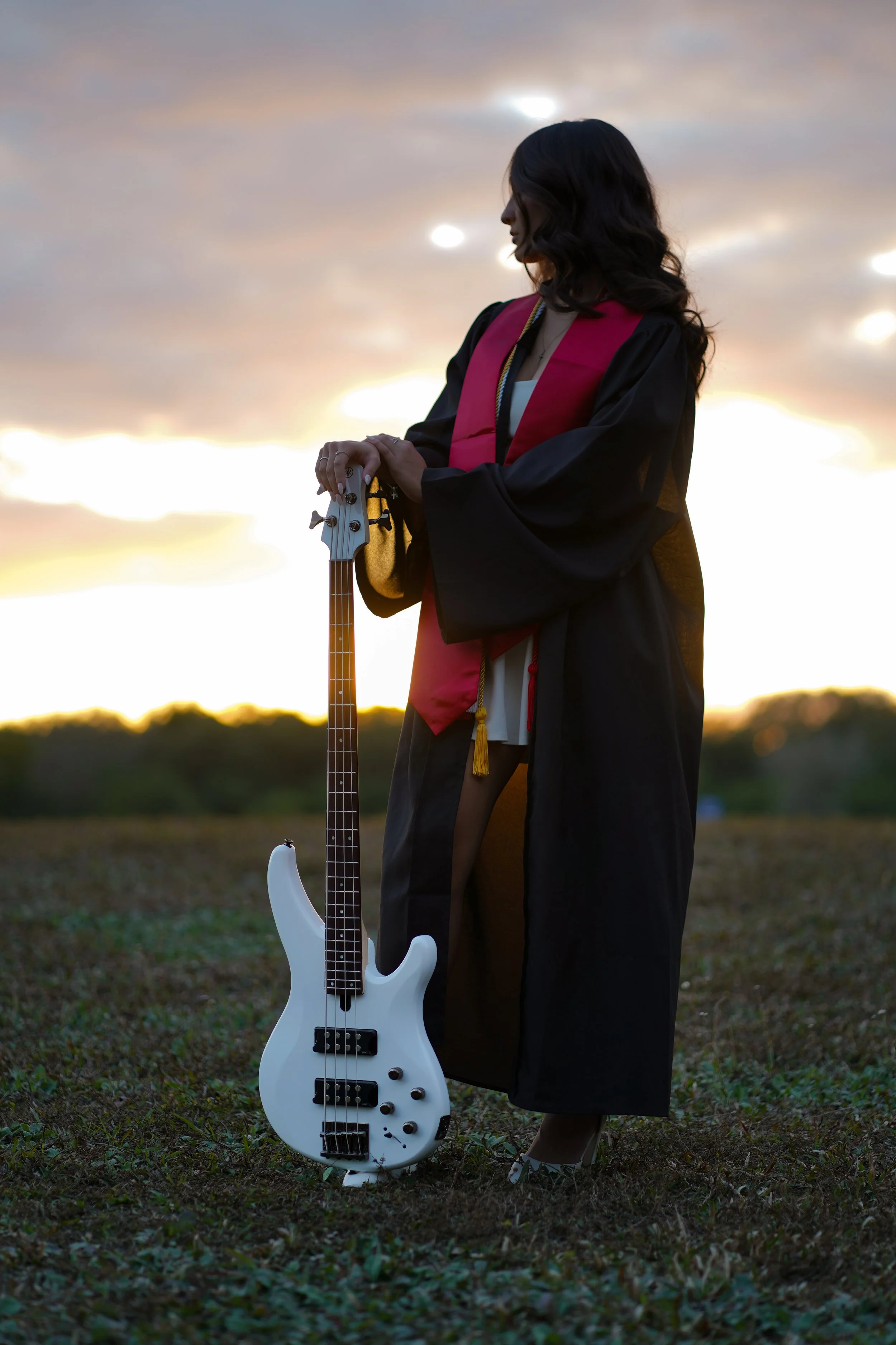 A woman in a graduation cap and gown holding a white electric bass guitar outdoors at sunset in Davie, Florida.