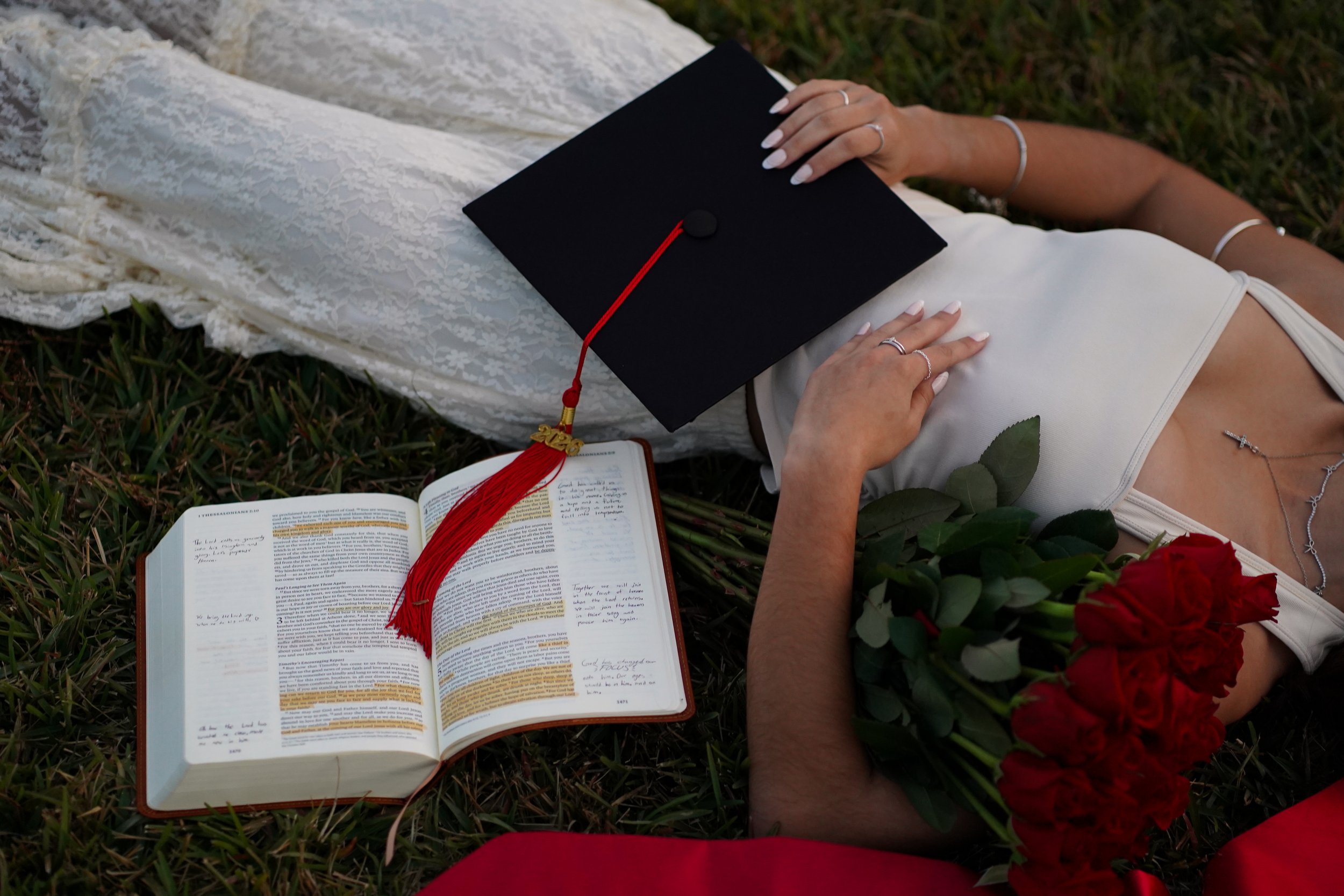A woman lying on the grass on her back, wearing a white dress and jewelry. She is holding a black graduation cap with a red tassel. An open book and a bouquet of red roses are nearby, indicating a graduation celebration in Florida.