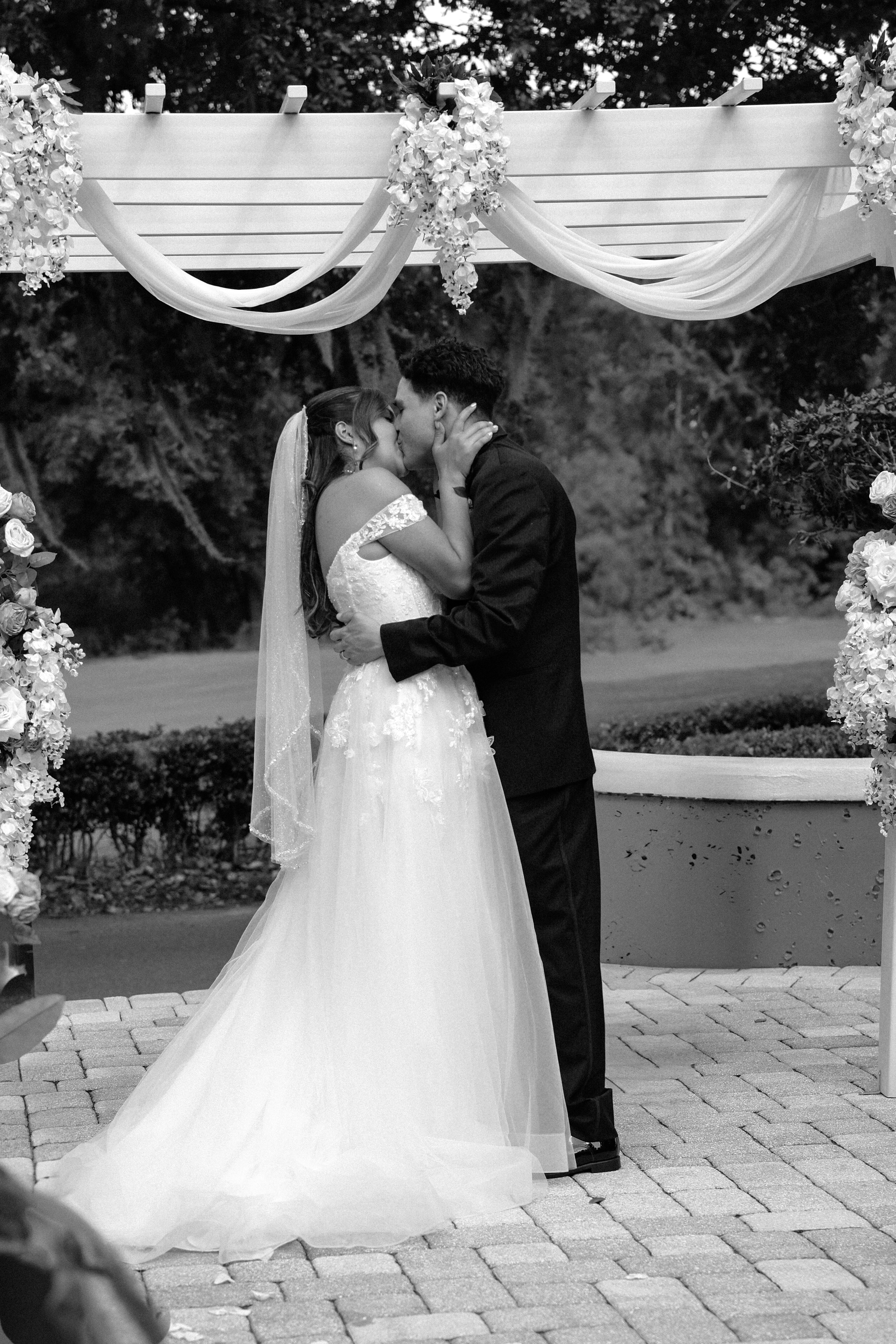 A couple sharing a kiss during their wedding ceremony outdoors, with floral decorations and draped fabric overhead, in black and white in Orlando, Florida.