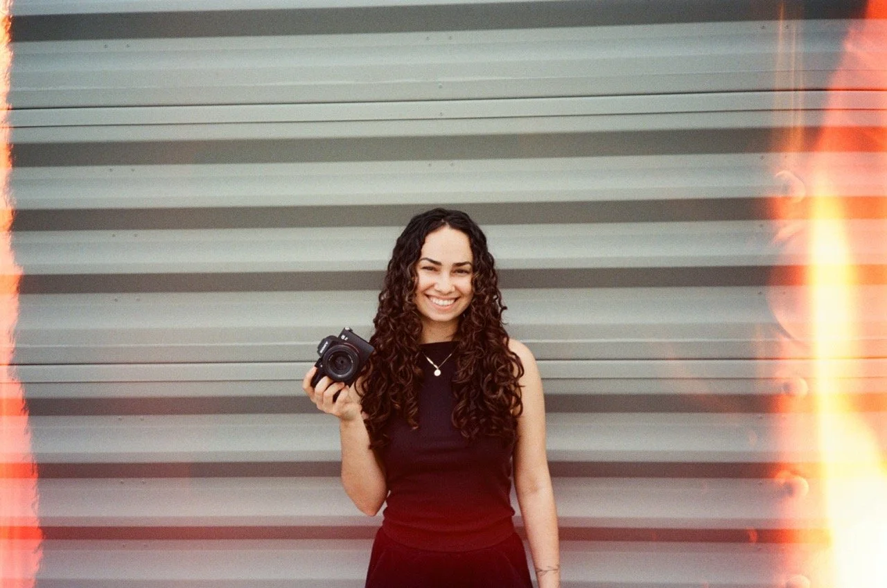 A woman with long curly hair smiling and holding a camera in front of a gray metal shutter door.