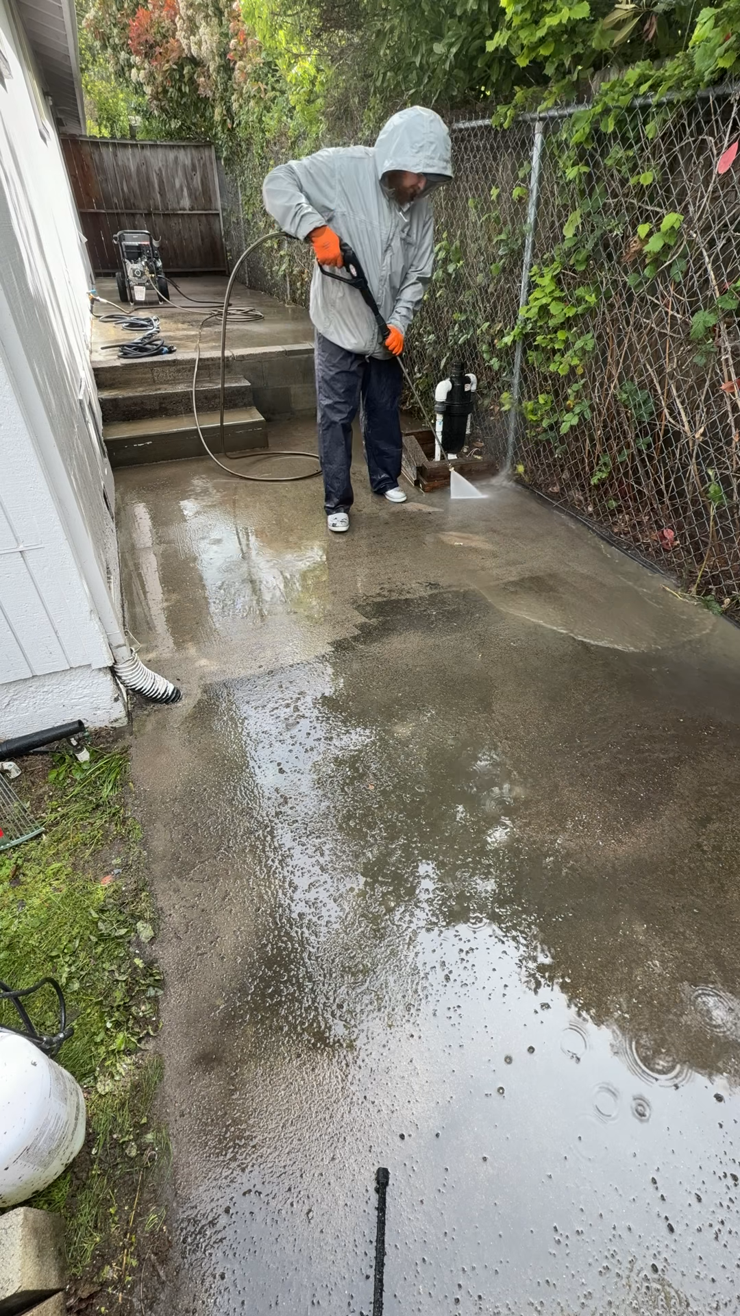 A person wearing a rain jacket and orange gloves power washing a concrete patio near a wooden fence and a house, with water and soap suds on the surface.