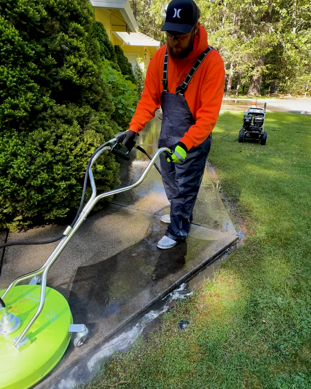 A man wearing an orange jacket, black pants, a black cap, and sunglasses is pressure washing a concrete sidewalk near a house, with a green pressure washer on the ground.