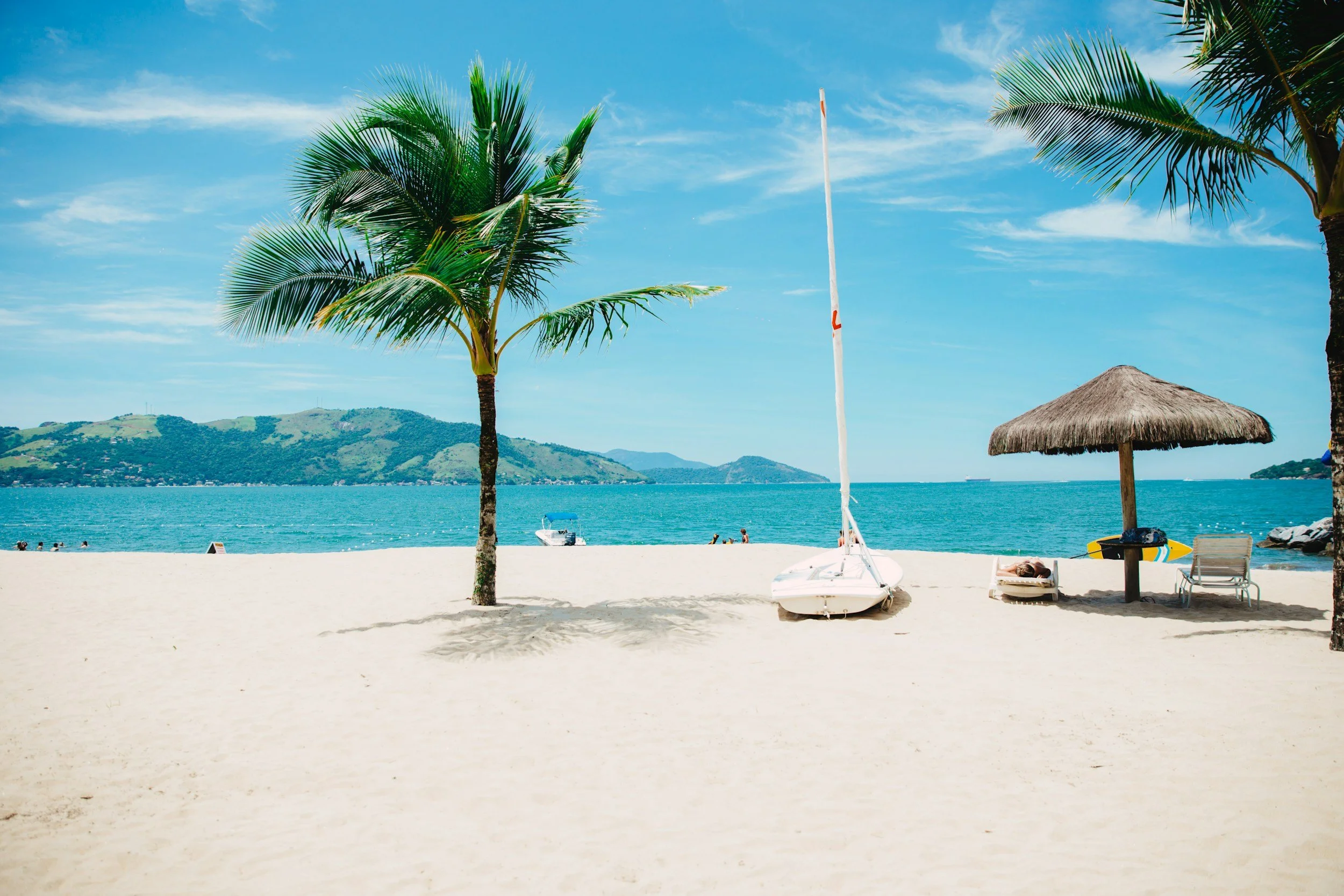 A sunny beach scene with palm trees, a sailboat on the sand, a thatched umbrella, lounge chairs, and a view of the ocean with hills in the distance.