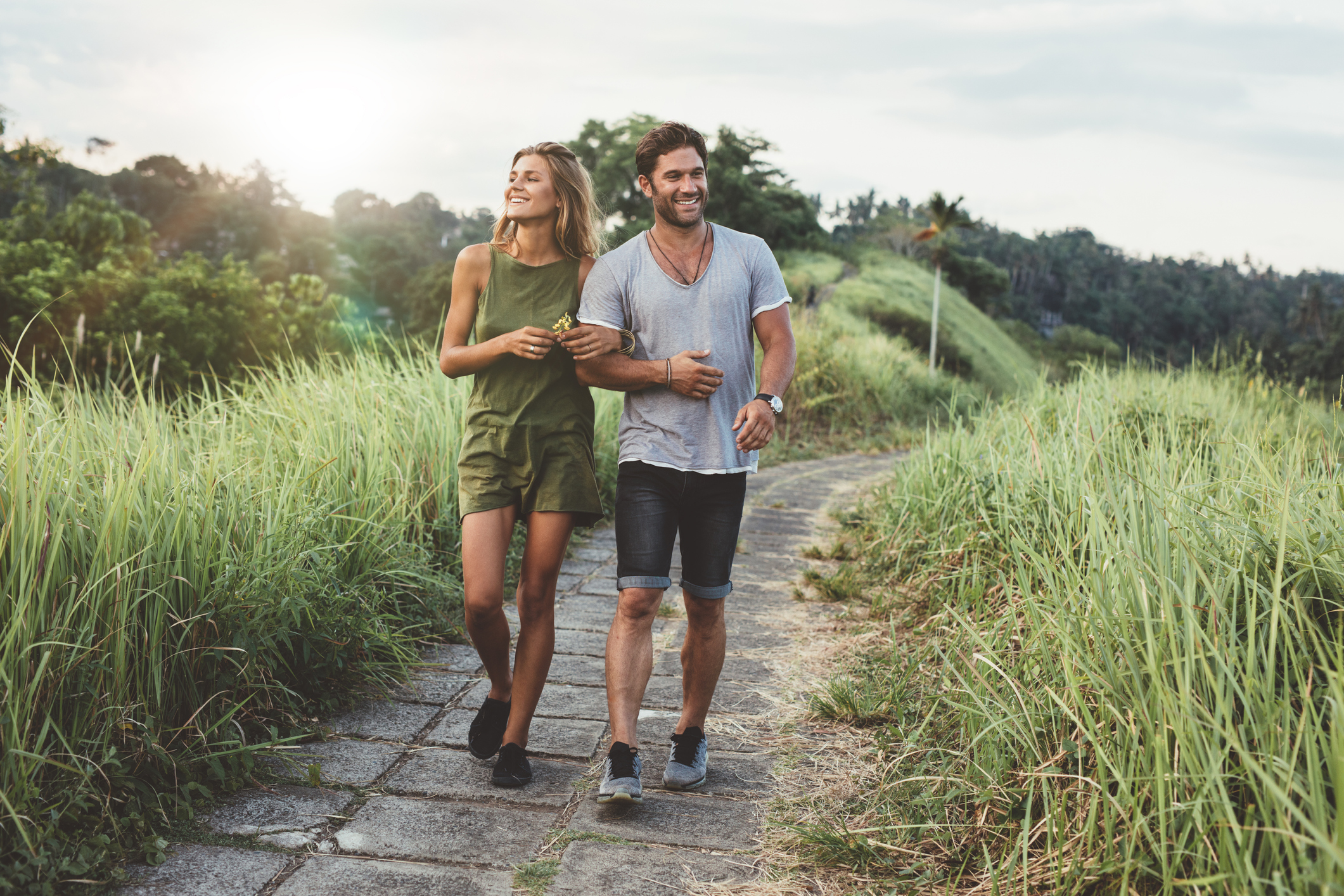 A young man and woman walking on a stone path through grassy hills, enjoying a sunny day outdoors.