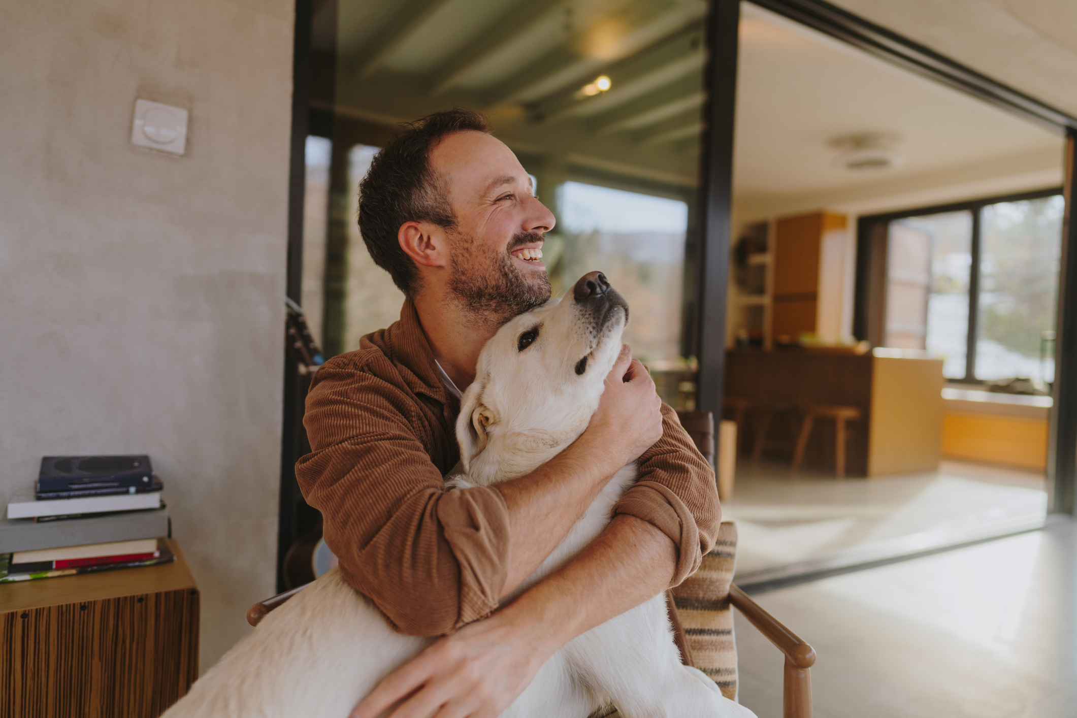 A man with a beard smiling and hugging a large white and tan dog indoors with large windows and modern decor.