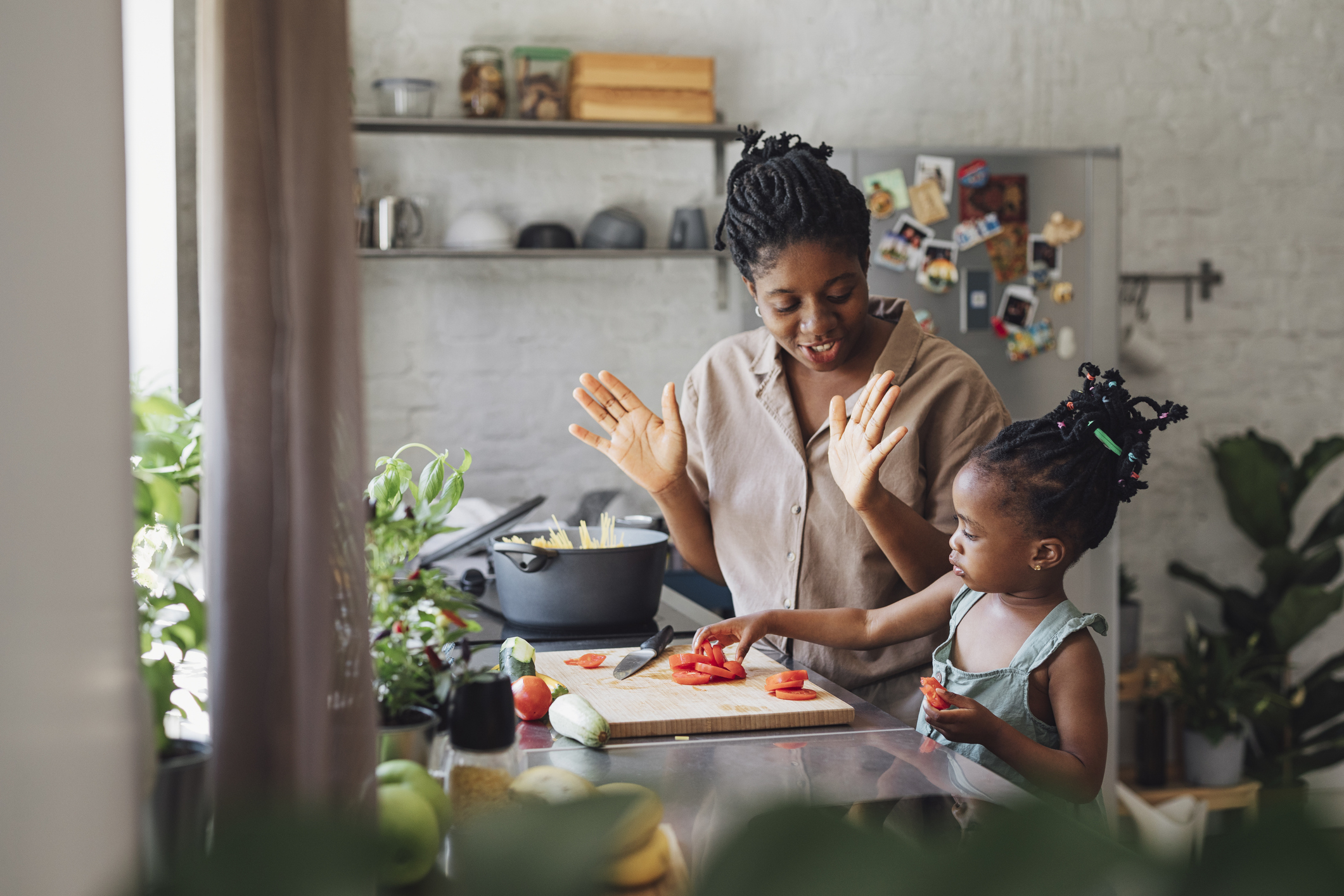 A woman and a young girl cooking together in a modern kitchen. The woman is smiling and engaging with the girl, who is cutting red bell peppers on a wooden cutting board. The kitchen is decorated with plants, photographs, and kitchenware.