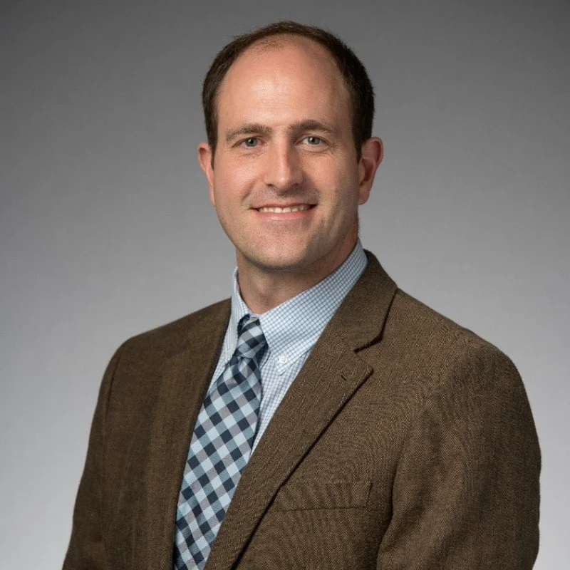 Professional headshot of a man wearing a brown blazer, light blue checkered shirt, and patterned tie, smiling against a gray background.