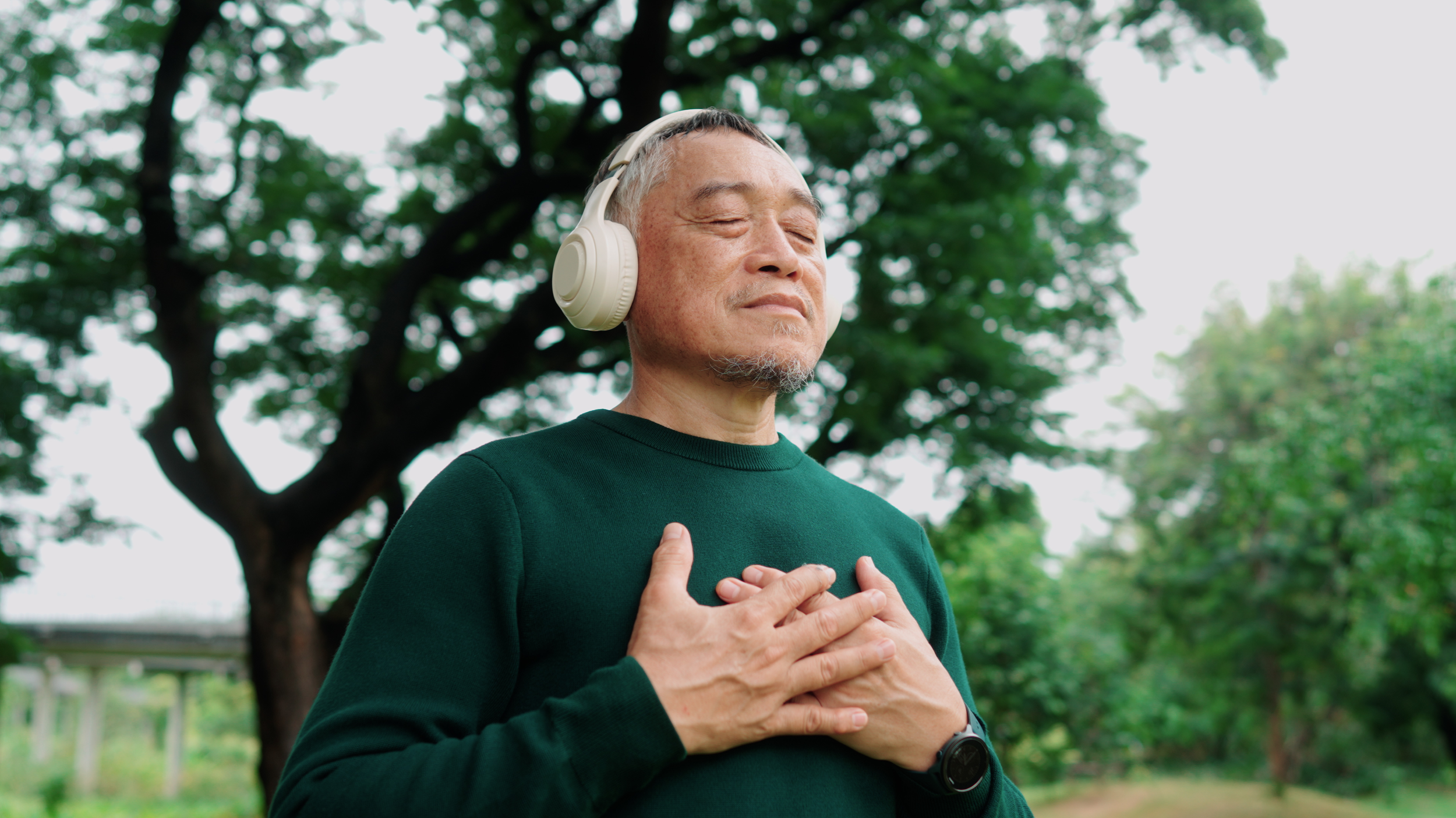 An elderly man with gray hair and a beard, wearing a green sweater and white headphones, stands outdoors with eyes closed and hands on his chest, surrounded by greenery and trees.